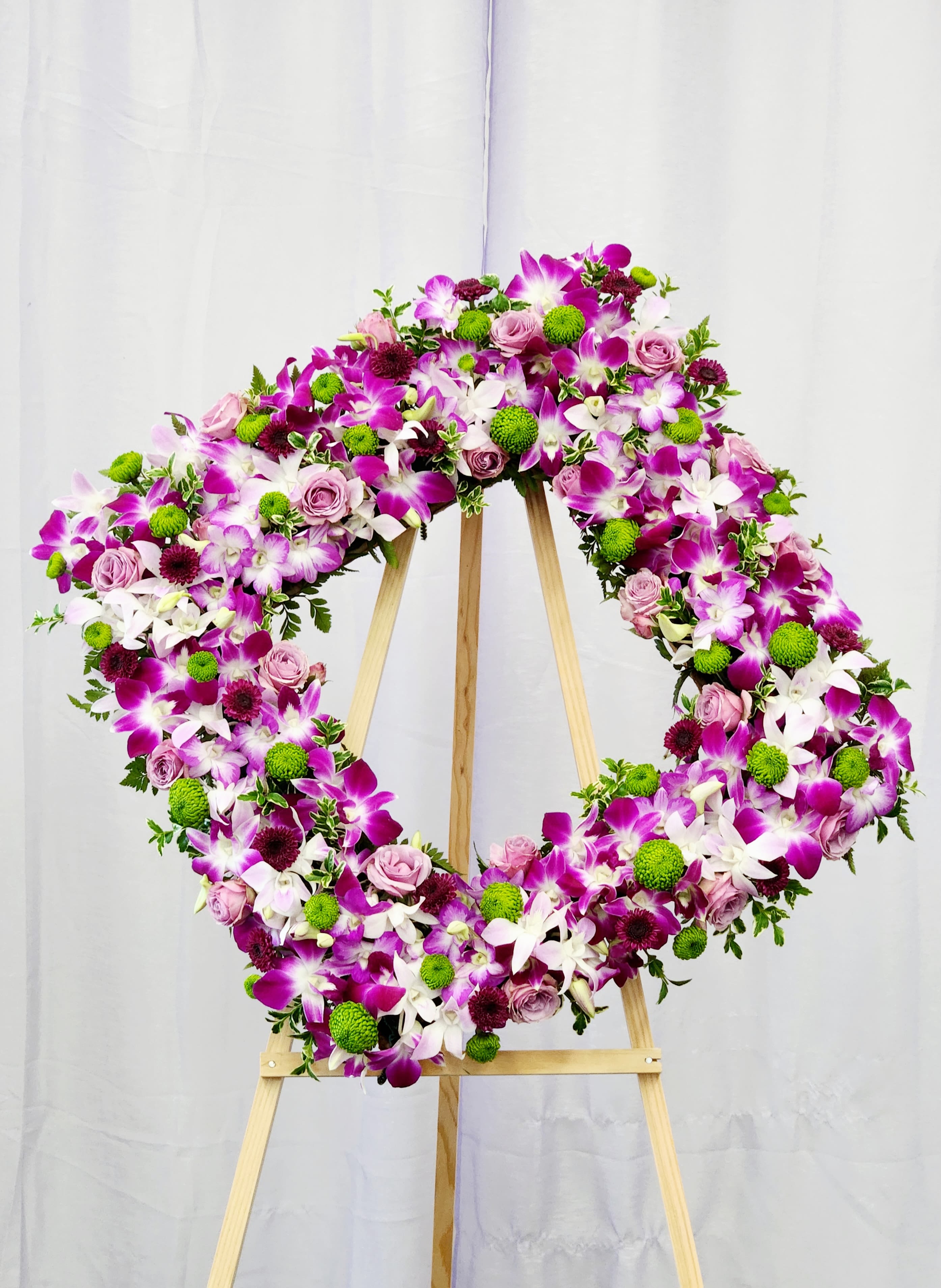 Orchid Picture Frame - A lush picture frame made of fresh vivid purple and white orchids, soft pink roses, and bright green chrysanthemum pompons presented on a sturdy wooden easel creates an immediate, ceremonial impression-ideal for a memorial tribute or a formal wedding display. The circular (and also available in a heart-shaped) silhouette reads as both comforting and celebratory, the kind of arrangement that helps the person you're honoring feel seen and remembered.  
