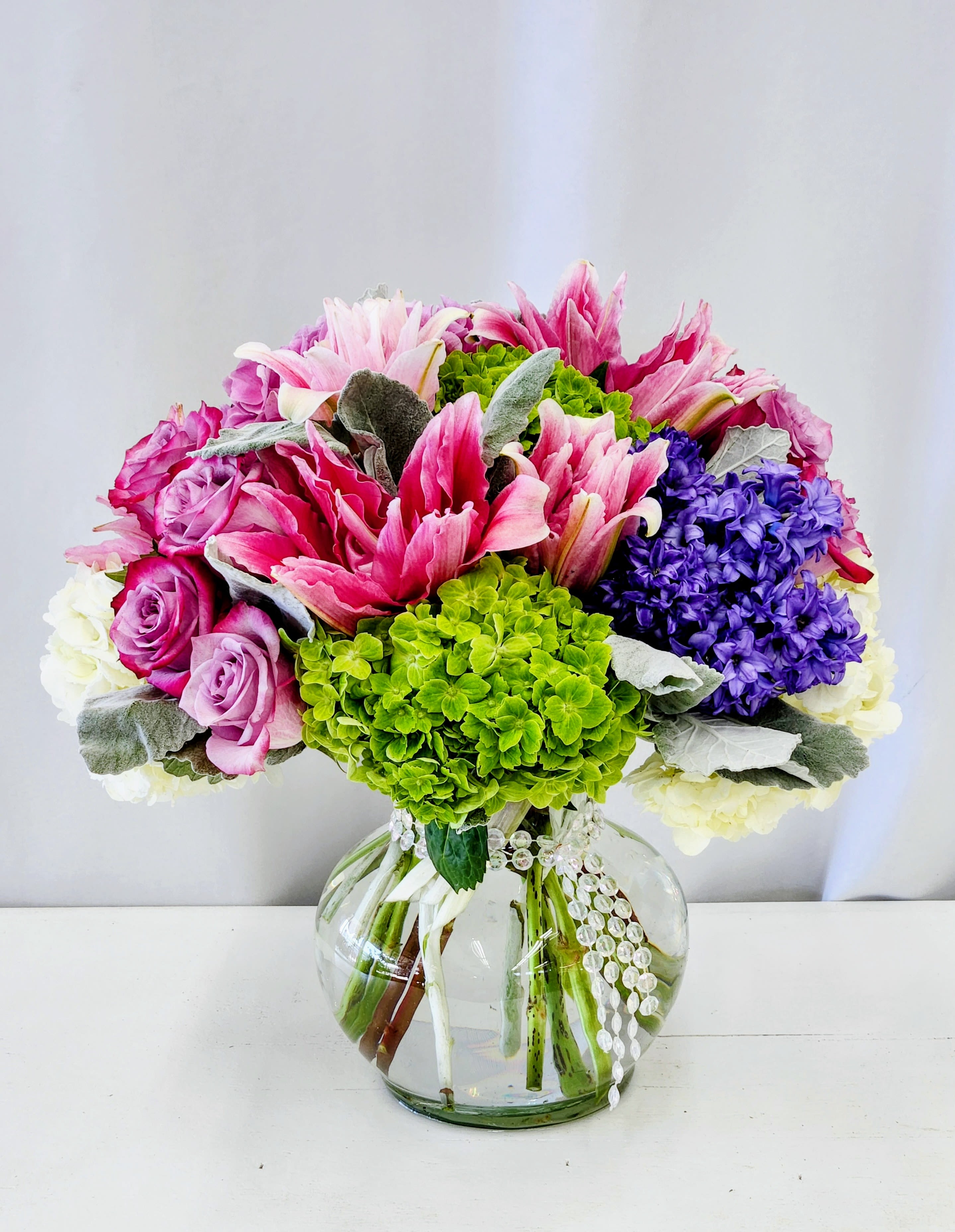 Harmony - Bright pink lilies and a mix of pink and lavender roses create an immediate burst of color in this celebratory flower arrangement-perfect as a birthday bouquet, anniversary flowers, or a heartfelt Mother's Day gift. The rounded glass vase and decorative beaded ribbon give it a polished, gift-ready look that makes the person you're thinking of feel genuinely celebrated the moment it's set down.  The arrangement pairs vivid green hydrangeas with clusters of purple blooms on one side, while pale white flowers and silvery foliage soften the edges for a balanced, layered texture. The composition is full and symmetrical, with lush stems arranged for an even, long-lasting silhouette. Delivered in a clear glass vase, it's an everyday-keepsake that reads cheerful and refined without being fussy.  