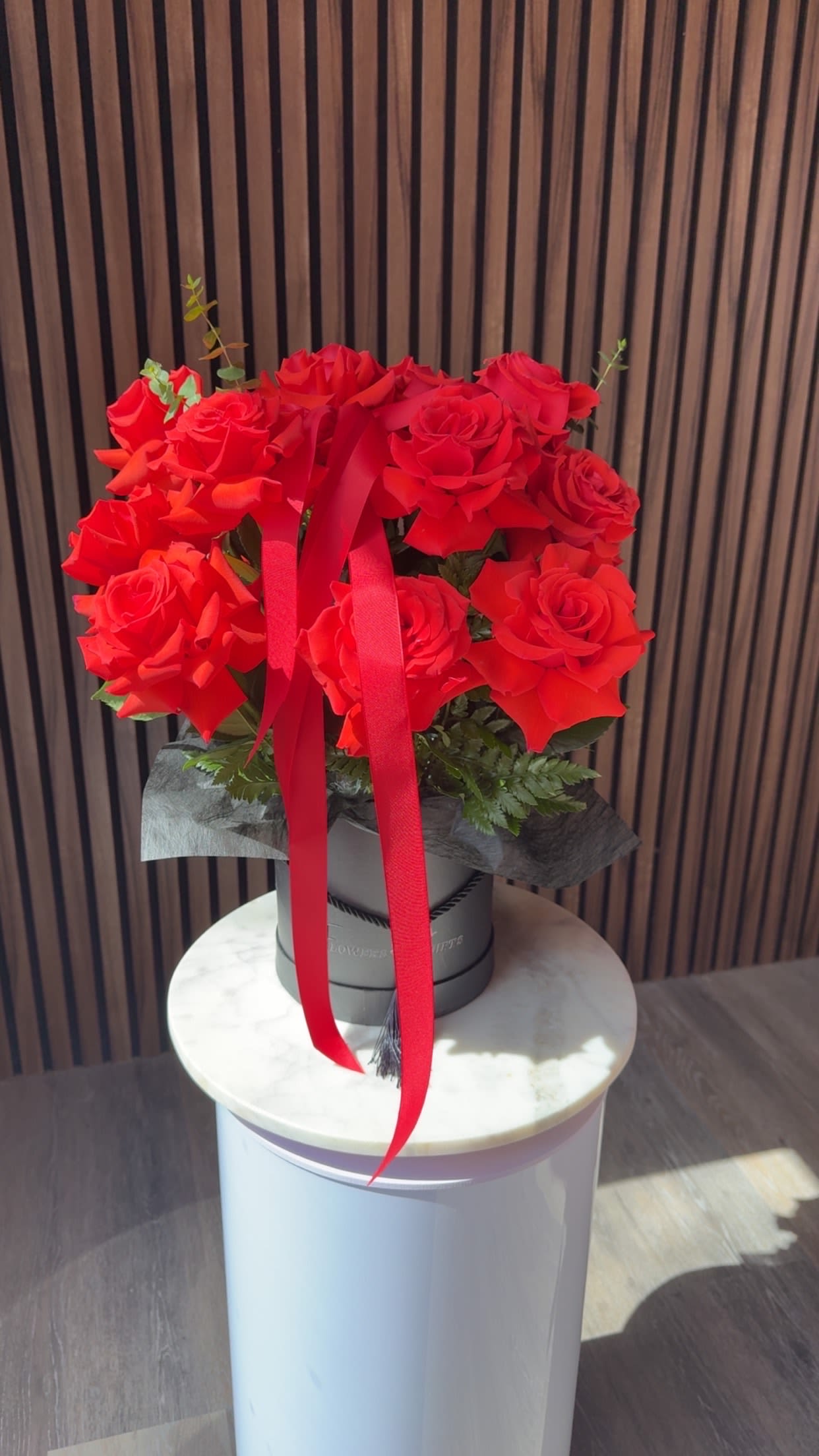 Nina Boxes - A lush cluster of vivid red roses shaped into a perfect,  rounded bouquet makes an immediate statement for Mother's Day, anniversaries, or any romantic occasion. The rich crimson blooms are arranged tight and low so the roses read as a single, luxurious mass-an unmistakable declaration of love that will make the person you're thinking of feel deeply cherished.  The roses sit in a sleek black hat box wrapped with a long red ribbon and a decorative outer wrap, the dark container creating a dramatic contrast that highlights each velvety petal. Small touches of greenery peek out beneath the blooms, adding texture and depth while keeping the focus on the flowers. The compact, hat-box presentation is easy to display on a table or bedside, and its formal, elegant look suits both classic and modern décor.  Perfect for marking anniversaries, romantic milestones, or saying "I'm thinking of you" on Valentine's Day, this arrangement sends a clear message of devotion. Order sizes are focused on the dense, rounded presentation. Please note care is simple: keep out of direct heat, refresh water if the floral foam is exposed, and enjoy the roses at peak bloom for several days.
