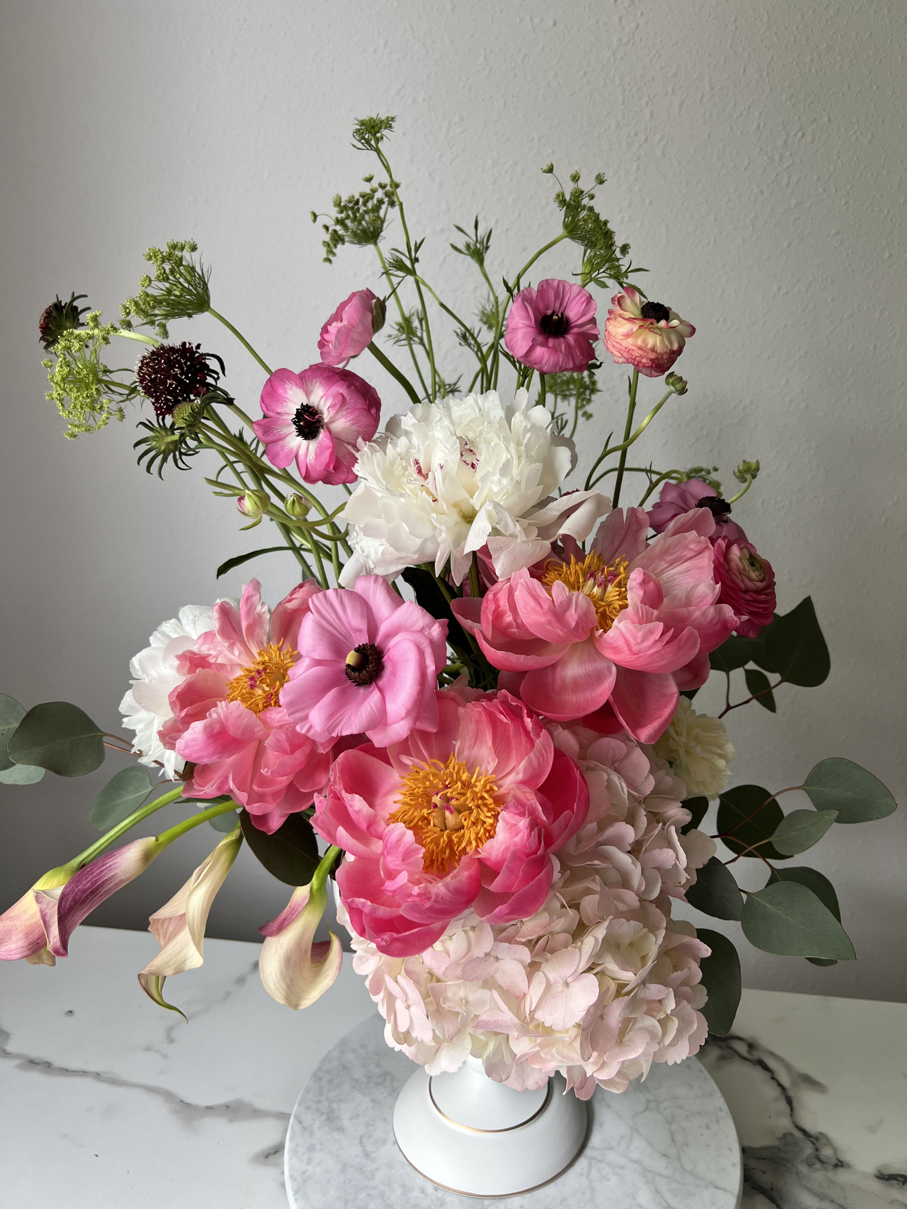 Blush Muse Bouquet - Soft, garden-fresh peonies set the tone for this Blush Muse bouquet - a romantic flower arrangement built to make the person you're thinking of feel truly seen. Perfect for Mother's Day, birthdays, anniversaries, or a just-because surprise, the mix of pink and white peonies with pink anemone-like blooms and airy green stems reads like a gentle, heartfelt embrace that says "you matter."  This vase arrangement pairs pale pink hydrangea and bright ranunculus with pale and mini calla lilies, lisianthus and delicate dianthus accents for layered texture and movement. Yellow-centered blooms and dark-centered anemones add contrast while lush green foliage lifts the composition; everything is arranged in a clean white ceramic vase for a refined, modern look. Sizes and exact varieties may vary with seasonal availability, but each bouquet is hand-designed to preserve the soft, romantic palette and garden-style silhouette.  Ideal for graduations, thank-you gifts, thinking-of-you moments, spring celebrations, weddings, or to welcome a new baby, the Blush Muse is created to convey warmth and celebration. Care is simple: keep water fresh, trim stems occasionally, and display out of direct heat to extend vase life; our florists will select the freshest blooms to deliver an arrangement that delights on arrival.