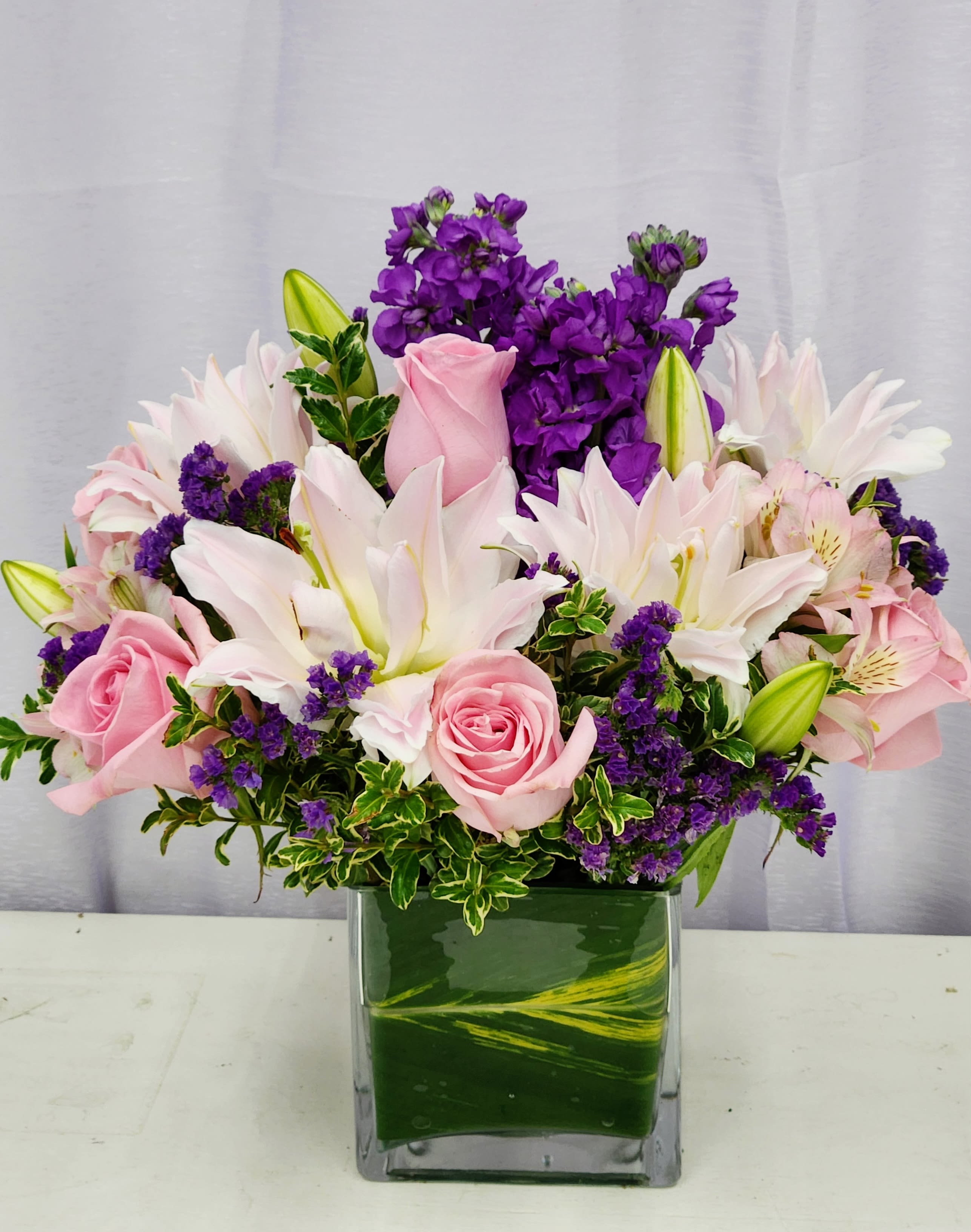 Purple and Pink Mist - Soft pink roses and large soft pink lilies mingle with clusters of vivid purple filler blooms in a rounded, garden-fresh composition-perfect as a birthday bouquet, anniversary arrangement, or a gentle sympathy display. The contrast of pale petals against saturated purple accents creates a romantic, slightly formal impression that will make the person you're thinking of feel comforted and truly seen.  
