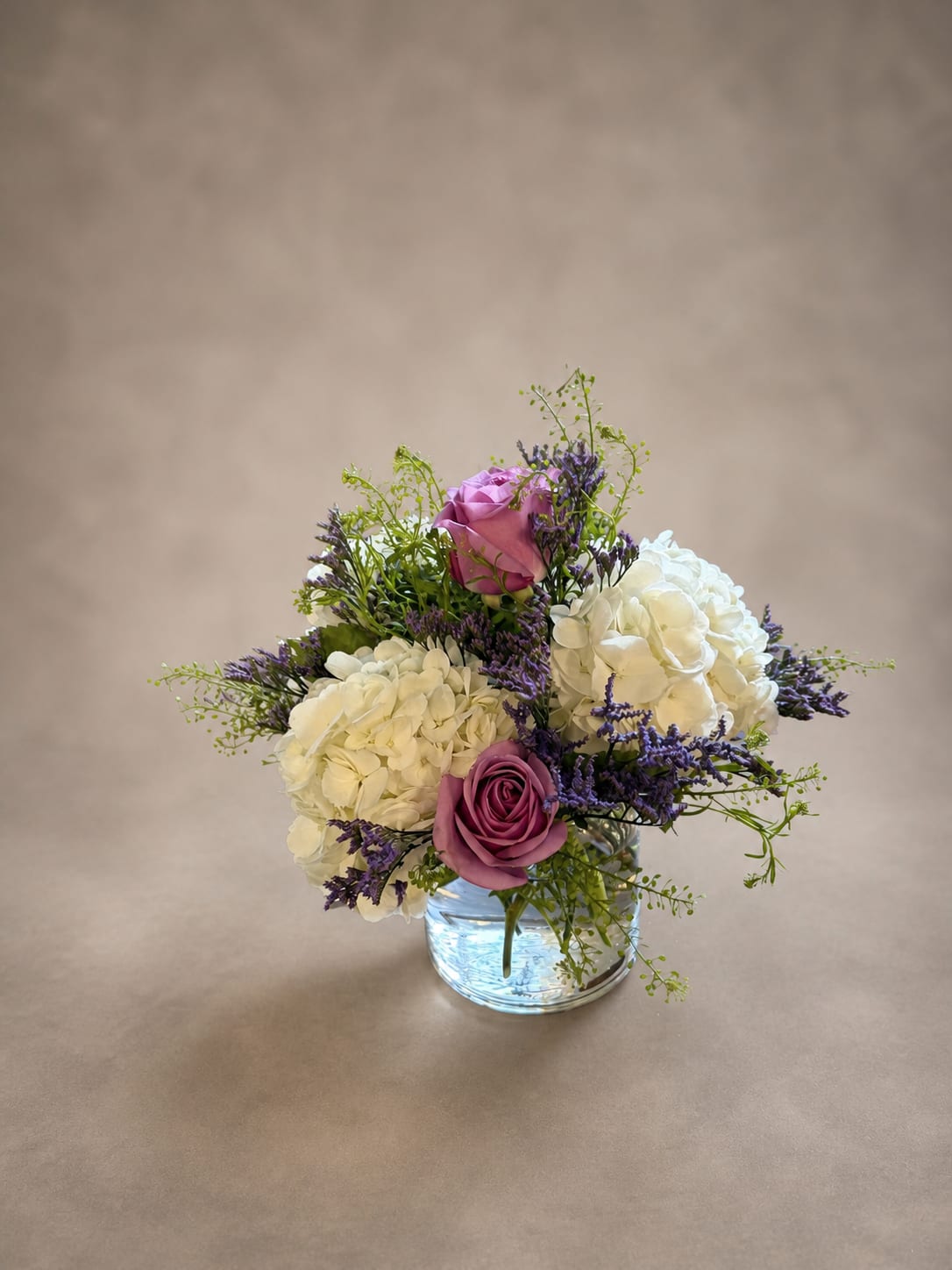 Lilac Hour - Some moments feel like the last light of a perfect day — soft, unhurried, and too beautiful to let go. The Lilac Hour captures that feeling in bloom. Lush white hydrangea cluster together like quiet memories, while purple roses open slowly at their own pace. Sprigs of lavender and delicate green filler dance in between, giving the arrangement an effortless, just-gathered feeling. Tucked into a simple glass vessel, this is a centerpiece that doesn't demand attention — it simply earns it. Perfect for weddings, intimate dinners, or anyone who deserves a little tenderness.