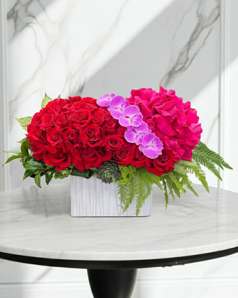 Crimson Luxe - A dramatic, romantic statement for anniversaries or Valentine's Day, this flower arrangement puts rich red roses and bright pink roses center stage so the person you're celebrating feels adored. The left side's dense mass of crimson roses balances the vibrant pinks on the right, while a sweep of purple Phalaenopsis orchids arcs across the middle to add sculptural contrast and a touch of elegance.  Set in a sleek white rectangular ceramic vase, the composition is full and highly saturated-deep reds, vivid pinks, soft white accents, and jewel-toned orchids create a luxurious, modern look. Fern fronds and lush greenery nestle around the base to soften the lines and give the piece a garden-fresh texture; the rounded, compact profile makes it ideal for a dining table centerpiece or bedside surprise.  Perfect for romantic milestones, celebratory gifting, or any moment you want them to feel truly seen, this arrangement arrives ready to display in its ceramic vessel. Keep flowers in fresh water, out of direct heat and sunlight, and replace water every couple of days to enjoy peak bloom longevity.