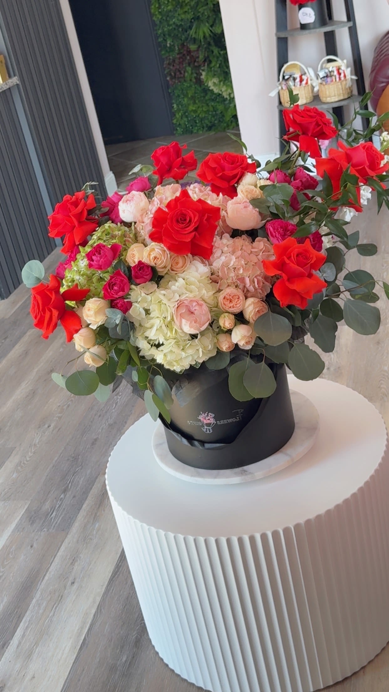 Garden Box - A lush, high-impact romantic bouquet designed to make the recipient feel deeply celebrated - presented in a sleek black hat box, this large mixed bouquet is perfect for a birthday, anniversary, Valentine's Day, or any special occasion when you want to say "I love you" in full color. Vivid red roses frame the arrangement and create an immediate, passionate impression, while blush and cream spray roses add soft, intimate highlights that balance the drama and keep the overall look tender and heartfelt.  At the center, a generous cloud of pale white hydrangeas gives the bouquet volume and a soft, tactile contrast to the more structured roses. Rounded eucalyptus foliage and cool green accents soften edges and add an organic, garden-fresh feel. The black hat box and coordinating black bouquet wrap make the reds and pinks pop for a polished, upscale presentation that reads romantic and celebratory.  This is the kind of gift that makes someone feel truly seen-romantic enough for an anniversary or Valentine's, joyful enough for a big birthday or congratulations. Large and abundant by design; please note the presentation comes in a black hat box (or with black wrap as shown) and is crafted to maintain its shape-keep blooms in a cool spot away from direct heat and refresh water as recommended for vase-flower care if you transfer them.
