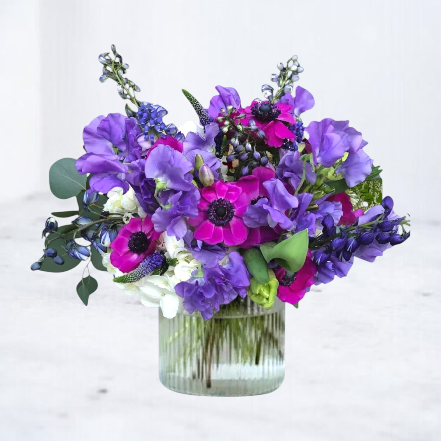 Afterparty - Afterparty is a bold purple and magenta flower arrangement designed with vibrant anemones, lavender sweet peas, delphinium, veronica, lisianthus, and premium seasonal blooms in a glass vase. Rich jewel tones and airy movement create a dramatic editorial floral design that feels energetic, artistic, and unforgettable.  Designed for birthdays, celebrations, statement gifting, modern sympathy flowers, luxury everyday flowers, and elevated event styling, Afterparty blends vivid purple textures with striking pink focal blooms for a contemporary arrangement that stands out from traditional florals.  Handcrafted with premium seasonal flowers and available for same-day flower delivery in Magnolia, TX and surrounding areas including The Woodlands, Montgomery, Conroe, Spring, Tomball, and Lake Conroe communities.