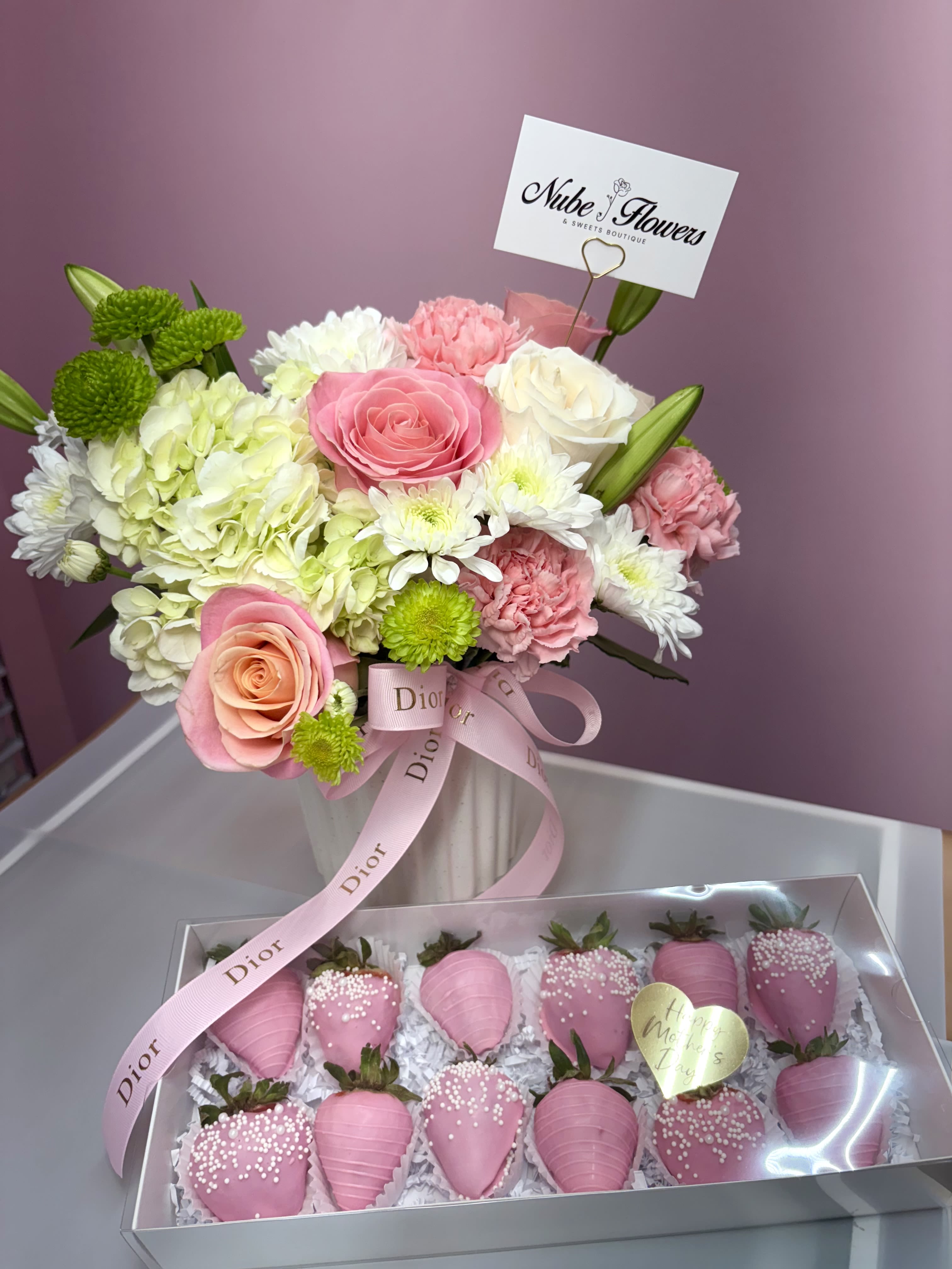 Blooms and Berries For Mom - Soft pink and white roses mingle with pillow-like hydrangeas and ruffled carnations in a compact, romantic bouquet that feels like a warm hug for Mom - perfect as a Mother's Day surprise or a thoughtful gift for birthdays and anniversaries. The mix of peachy tones, bright green button mums, and unopened lily buds gives the arrangement a fresh, playful lift so the recipient will feel cherished and celebrated the moment they open it.  Arranged in a classic white vase and tied with a pale pink ribbon, the bouquet pairs with a boxed set of pink-dipped strawberries presented under a clear lid for an extra-sweet touch. Textural contrast comes from chrysanthemums and the glossy berries while a heart-shaped tag and branded floral pick card finish the luxe presentation. Sized to sit beautifully on a bedside table or dining centerpiece, the arrangement arrives ready to display - recommend trimming stems and changing water every two days for best freshness.