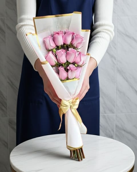Pink Wrapped Bouquet - A compact cluster of soft pink roses makes this wrapped bouquet an immediate expression of affection-perfect as a birthday bouquet, anniversary roses, or a simple "thinking of you" gift. The rounded, hand-tied form gives a fresh, intimate feel so the person you're honoring will feel truly seen and cherished the moment they unwrap it.  The roses are arranged tightly in a gentle pink palette accented by touches of white and yellow, then wrapped in clean white decorative paper with subtle gold edging and tied with a pale yellow satin ribbon. The finish is polished and gift-ready, with a tactile mix of velvety petals, crisp paper, and smooth ribbon that reads romantic without fuss.  Ideal for everyday gifting or more personal celebrations, this bouquet arrives ready to present-no vase required. To keep the roses looking their best, trim stems at an angle and change water daily if transferred to a vase; otherwise let the recipient enjoy the ready-to-give presentation as shown.
