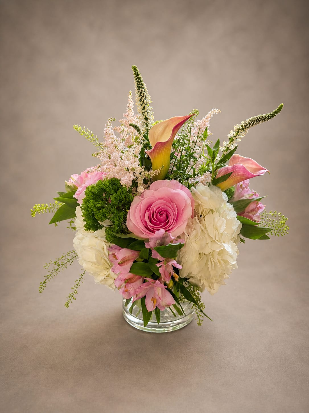 Sunday morning - There's a particular kind of beauty that doesn't try — it simply is. "Sunday Morning" is that feeling made tangible. A full pink rose sits at the heart of the arrangement like a quiet confidence, surrounded by soft white hydrangea, delicate alstroemeria, and feathery astilbe that catches the light like it was made for it. Blush calla lilies rise gracefully above the rest, unhurried and elegant, while touches of green trachillium and wispy veronica add life and movement. Set in a low, rounded glass vase, this arrangement feels equally at home as a wedding centerpiece, a birthday gift, or simply on a table where someone deserves to feel loved on an ordinary day. Because the best mornings aren't grand — they're just full of the right things.