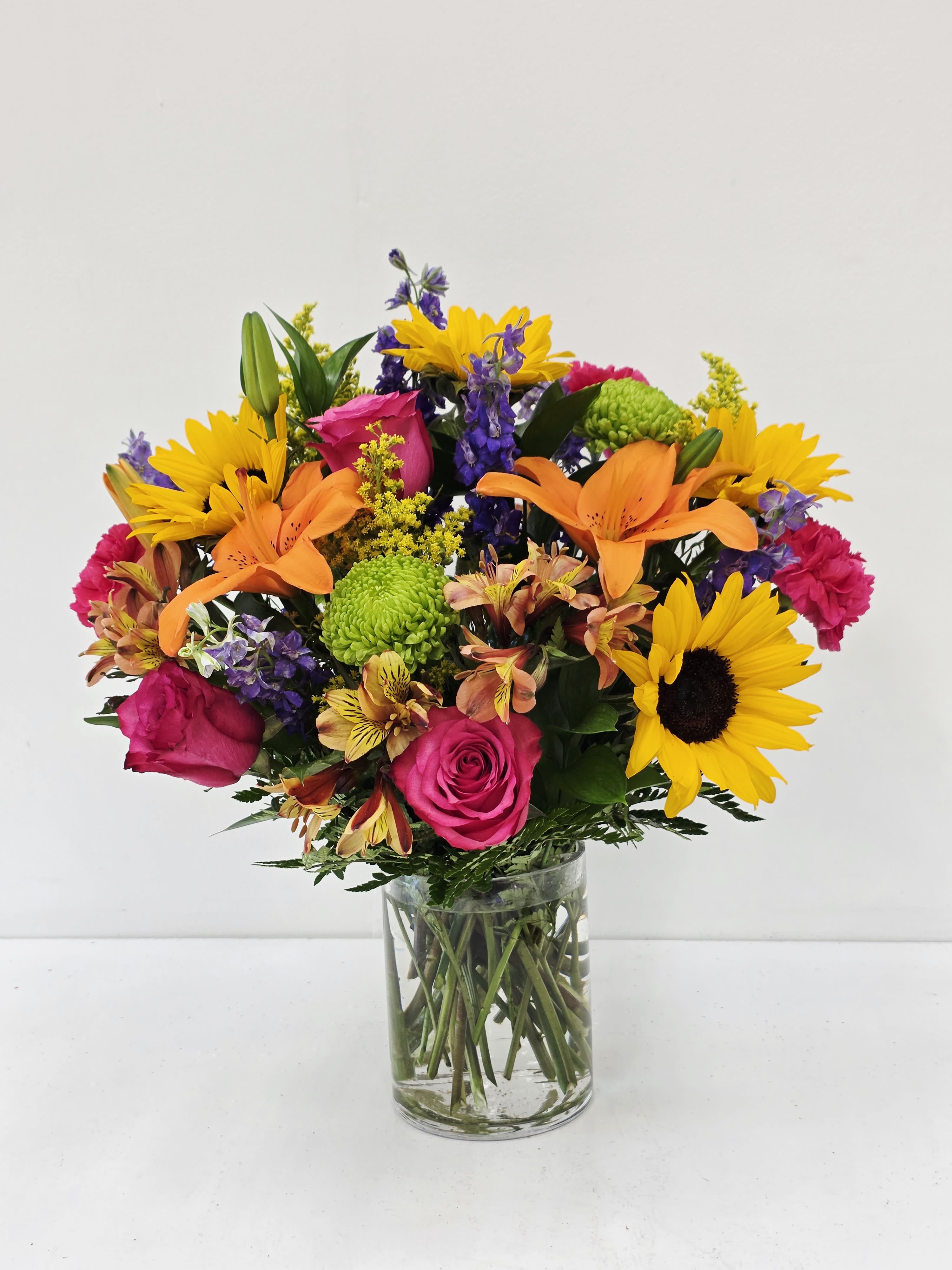 Fun in the Sunflowers - A burst of sunny warmth greets the recipient the moment they see this mixed color bouquet - vivid yellow sunflowers paired with orange lilies and hot pink roses create an energetic, tropical-tinged look perfect for a birthday bouquet, congratulations, or any cheerful everyday celebration. The clear glass vase shows off the stems and keeps the design feeling fresh and modern, so the person you're thinking of will immediately feel noticed and uplifted by the bright, confident color story.  
