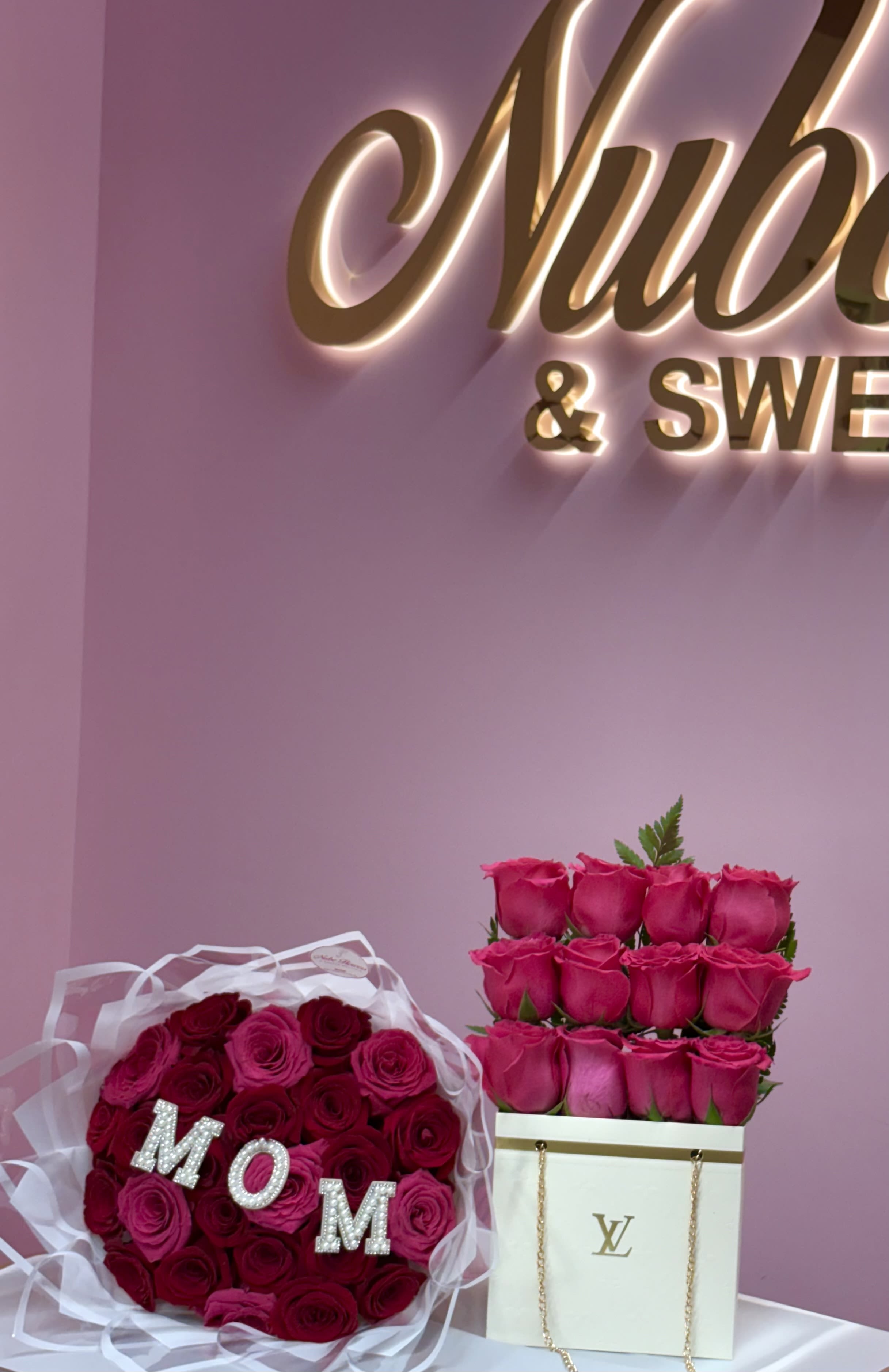 Mom Bundle  - Two matching rose arrangements that read "MOM" at a glance - a bold, feminine gift for Mother's Day or a special birthday. The Mom Bundle pairs a round bouquet of hot pink and deep red roses wrapped in sheer translucent material and finished with rhinestone letter picks spelling MOM, alongside a stacked display of pink roses in a white gift bag-style box with chain handles. Together they make a celebratory, luxury-feeling presentation that immediately says appreciation and love.  The roses show off saturated pinks and rich crimson tones against crisp white packaging for striking contrast; textures range from velvety petals to gleaming rhinestones and glossy chain handles. The set arrives ready to gift in a decorative gift bag and gift box, so the person you're honoring will feel caught up in the moment from the minute they open it. Ideal for the mom who loves bold color and thoughtful details, this bundle creates a memorable centerpiece and keepsake presentation she'll cherish.