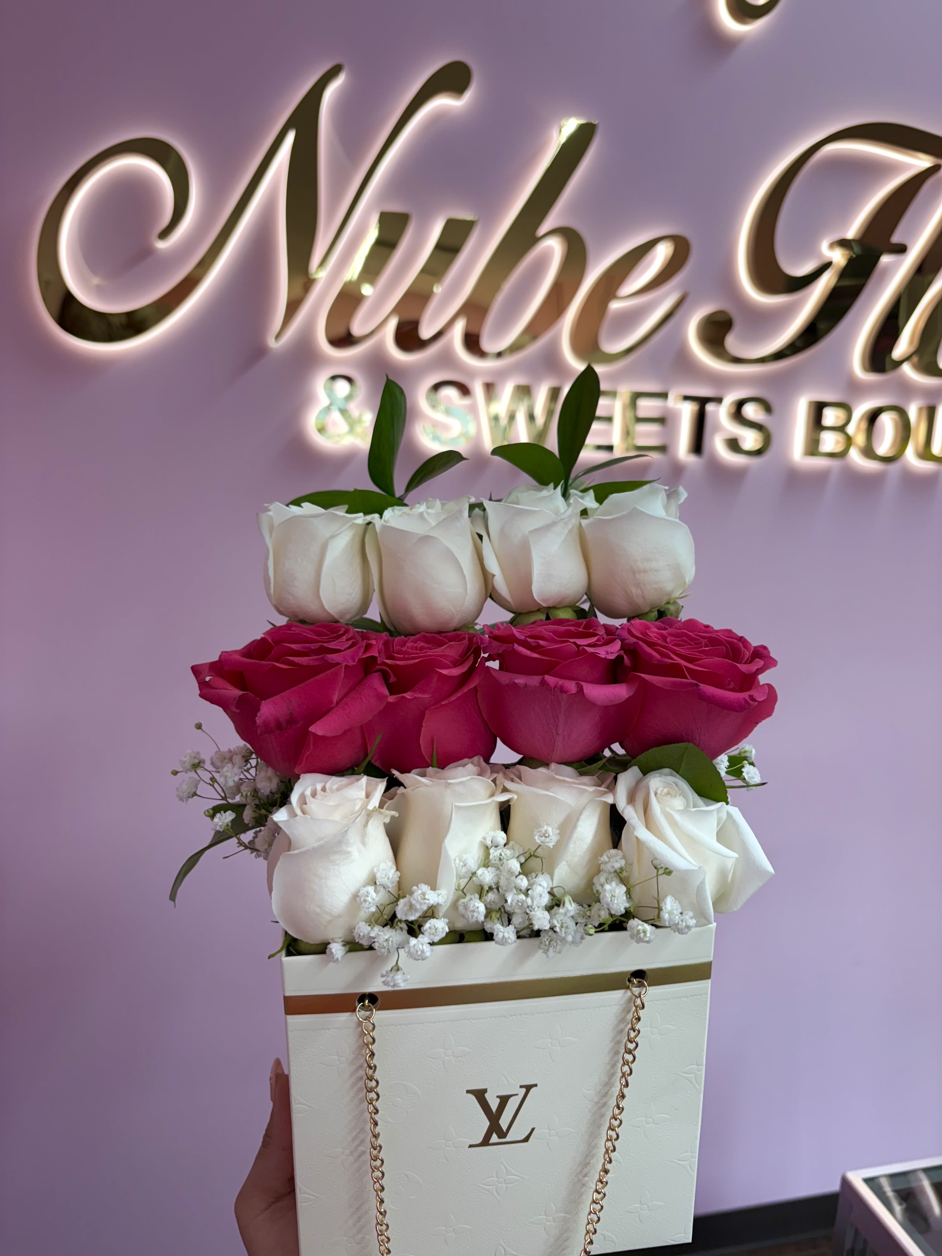 White LV floral bag  - Two-tiered roses in vivid pink and soft white create an immediate romantic statement-this flower arrangement in a white luxury gift bag is perfect as an anniversary bouquet, birthday surprise, or a heartfelt love-and-romance gesture. The compact, stacked composition reads like a polished present the moment she sees it, and the addition of delicate baby's breath brings an airy, tender contrast that makes the recipient feel truly celebrated.  Arranged inside a white luxury-style bag with gold chain handles, a metallic brand logo, and decorative ribbon, the bouquet looks as finished as a wrapped gift-no extra vase required. The arrangement pairs rich pink roses with creamy white roses and small clusters of baby's breath, accented by fresh greenery for texture and balance. Its tidy, two-tiered build keeps the display compact and travel-friendly while delivering high visual impact.  Ideal for marking anniversaries, birthdays, or any romantic milestone, this keepsake-style presentation sends a clear message of thoughtfulness and care. Hand-delivered in its branded gift bag, it's simple to present and easy for the recipient to enjoy; light trimming of stems and fresh water in a vase will extend the life of the roses if they choose to re-arrange them.