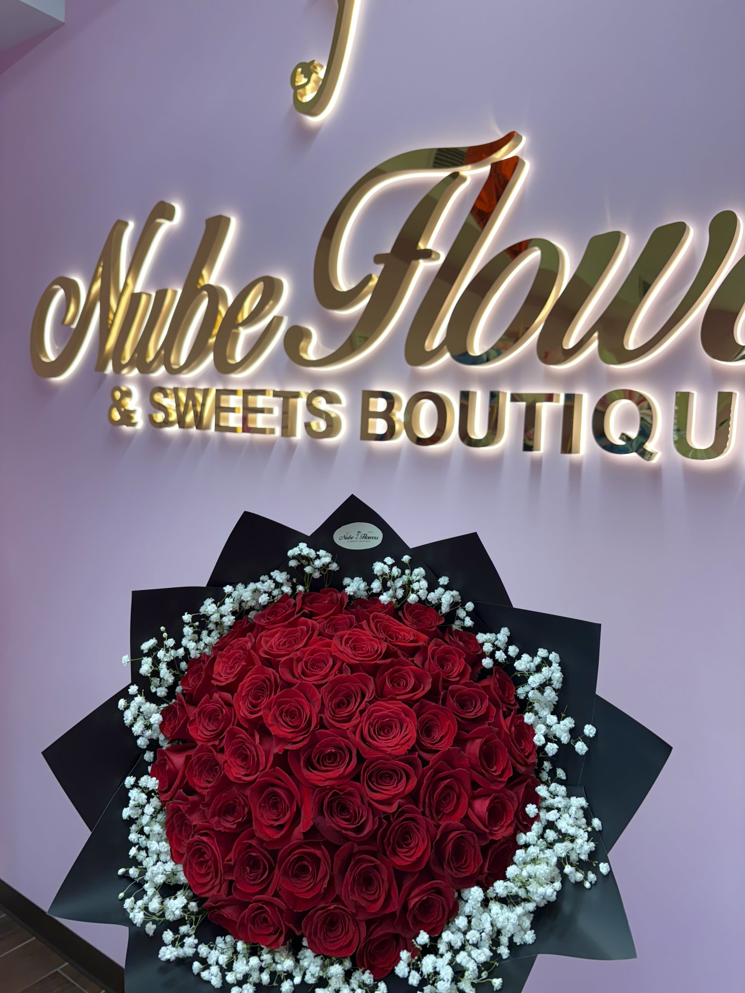 Nube Bouquet  - A dense, rounded cluster of deep red roses framed by a halo of delicate white baby's breath-this romantic bouquet makes a bold statement for an anniversary, Valentine's Day, or any moment you want to say "I love you" with dramatic flair. The contrast of passionate red and soft white against matte black wrap gives the arrangement a luxurious, formal presence that will make the person you're thinking of feel truly seen and adored.  Hand-tied and finished in a sleek black bouquet wrap, the arrangement's compact, rounded shape reads elegant and gift-ready straight from delivery. The roses form the visual heart while the baby's breath adds airy texture and a softly fragrant note; the black paper enhances color depth for a striking unboxing. Ideal for romantic celebrations or an upscale congratulatory gesture-order with standard sizing suitable for presentation; gently trim stems and refresh water promptly to extend vase life.