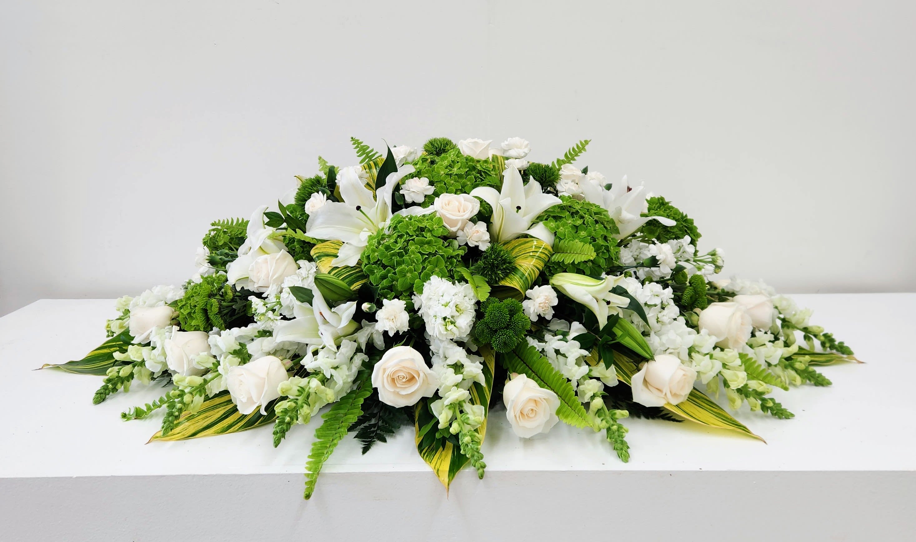 Memorable Soul - A calm, formal spread of white roses and lilies set against vivid green hydrangea makes this casket spray a quietly powerful choice for a sympathy arrangement or memorial centerpiece. The clean white-and-green palette and symmetrical, layered shape create a serene visual that helps the person you're honoring feel remembered and respected.  Dense clusters of white roses and lilies sit amid bright green hydrangea, accented with white filler blooms, trailing fern, and variegated tropical-style foliage for texture and contrast. The design is finished low and wide for a polished, table-level silhouette-ideal for a service program, memorial table, or as a dignified focal piece at a funeral home or celebration of life.  