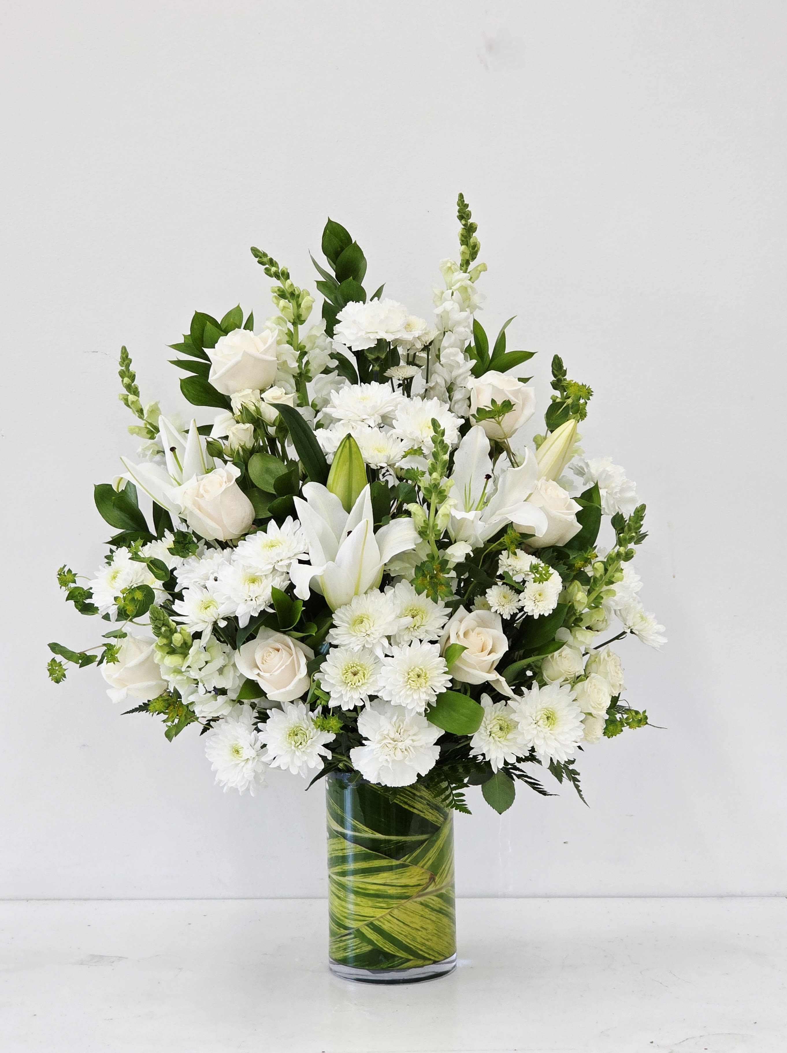 Purest Love - A serene cluster of white lilies, roses, chrysanthemums and tall snapdragon spikes creates an immediate sense of calm-the perfect sympathy arrangement or wedding centerpiece to express Purest Love. This design places the flowers in a soft white-and-green palette so the person you're honoring feels comforted and seen; it also works beautifully for anniversaries or to mark a quiet, heartfelt moment.  