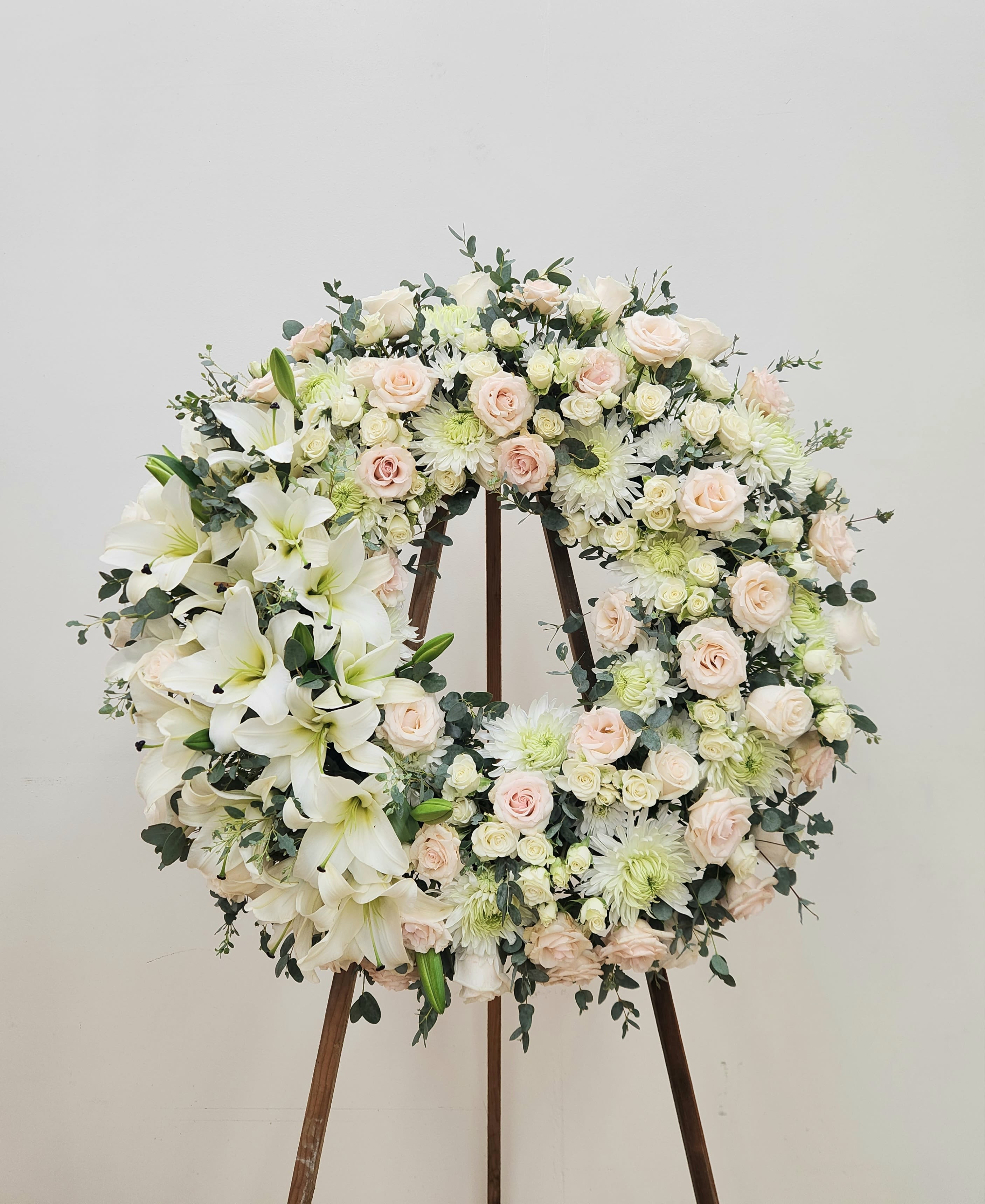 Sweet Farewell - A large circular wreath of white lilies, blush roses, spray roses and pale chrysanthemums creates a serene, elegant tribute perfect for sympathy or memorial services. Displayed on a wooden easel stand, this funeral wreath reads as calm and respectful-the kind of arrangement that helps the person you're honoring feel remembered and comforted.  Flowers are densely packed around the ring with airy eucalyptus-like greenery woven through for soft contrast, giving the bouquet a layered, textural look in white, cream, blush and pale green. The scale and traditional wreath shape present beautifully at a service or graveside; the restrained palette also makes this suitable for formal ceremonies or a wedding memorial when a quiet, refined touch is desired.