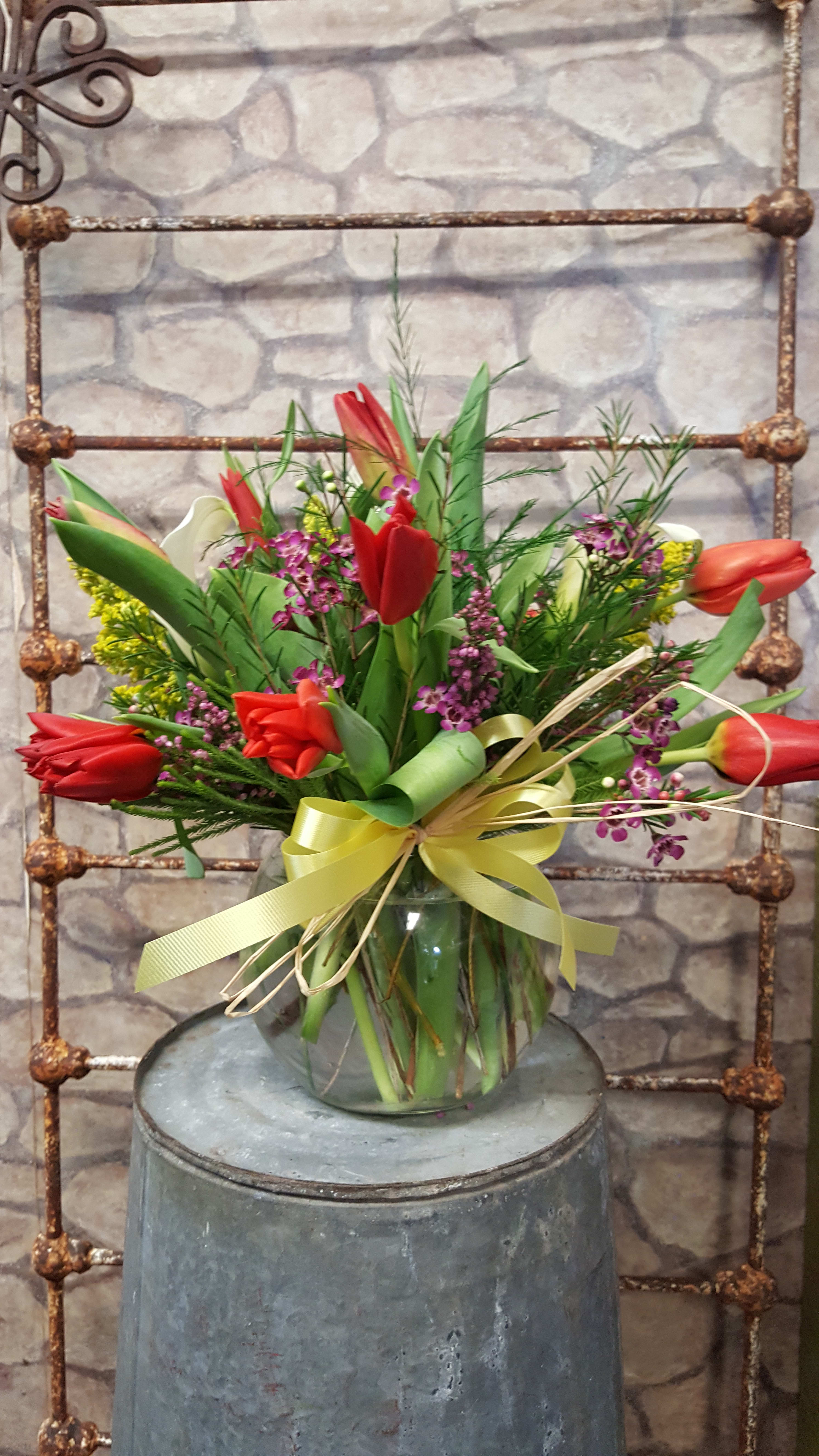 Mixed bouquet of red tulips and small purple flowers in a clear glass vase with yellow ribbon.