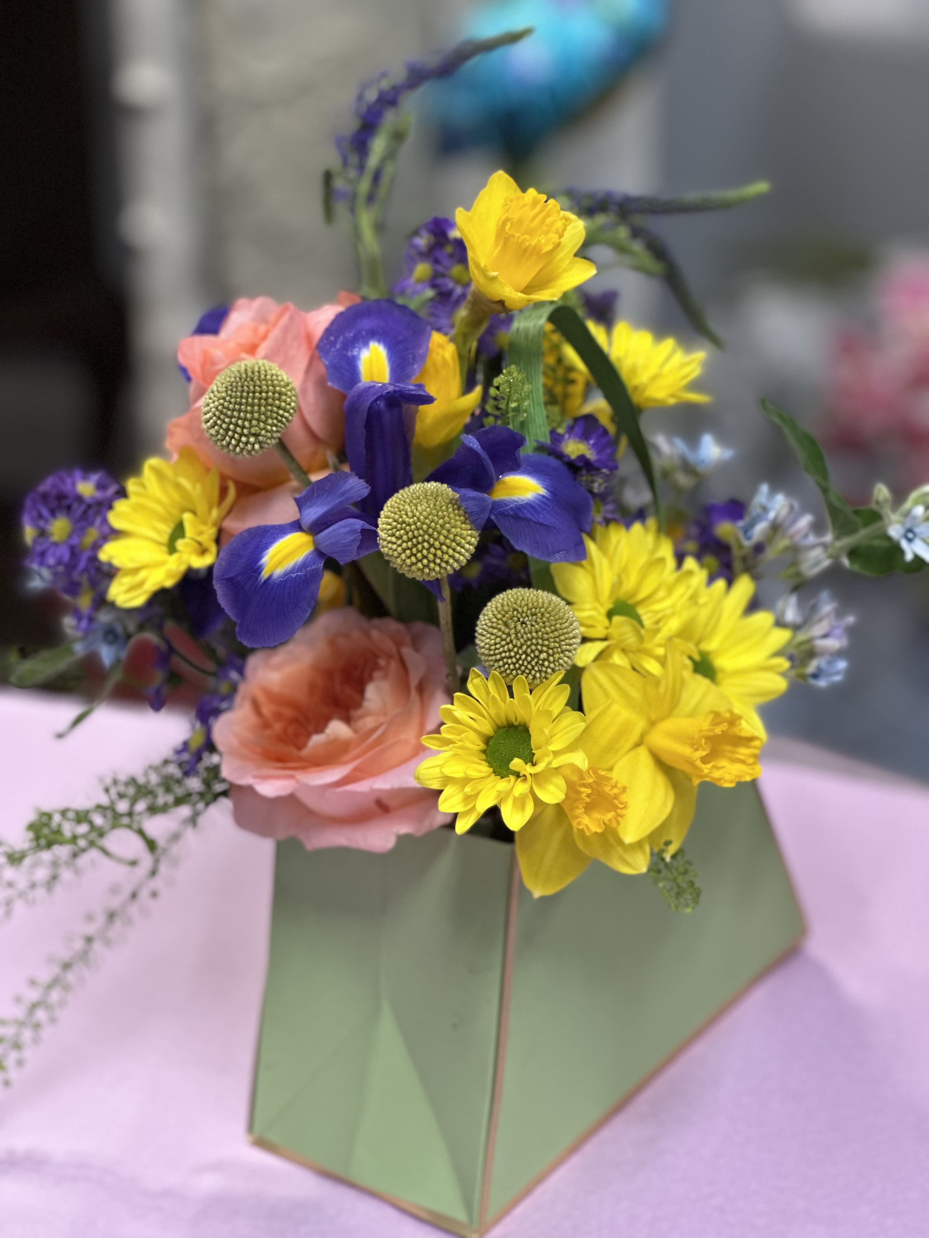 Mothers Day Purse  - Bright bursts of yellow daisies and deep purple iris pop against soft peach-pink and lavender roses in a compact, purse-style flower arrangement-an uplifting gift for Mother's Day, a spring birthday, or to celebrate love and friendship. The high-contrast mix feels garden-fresh and playful, the kind of bouquet that makes the person you're thinking of smile the moment they see it.  A hand-tied composition of daffodils, hydrangea accents, spray roses, daisies, iris and roses is finished with small green button-like accents for texture and a pale green gift bag with matching paper wrap for an effortless, ready-to-give presentation. Compact and modern in scale, it's an ideal pick for Spring, Women's Day, Best Friends Day, or when you want a bright, romantic gesture-no vase required.
