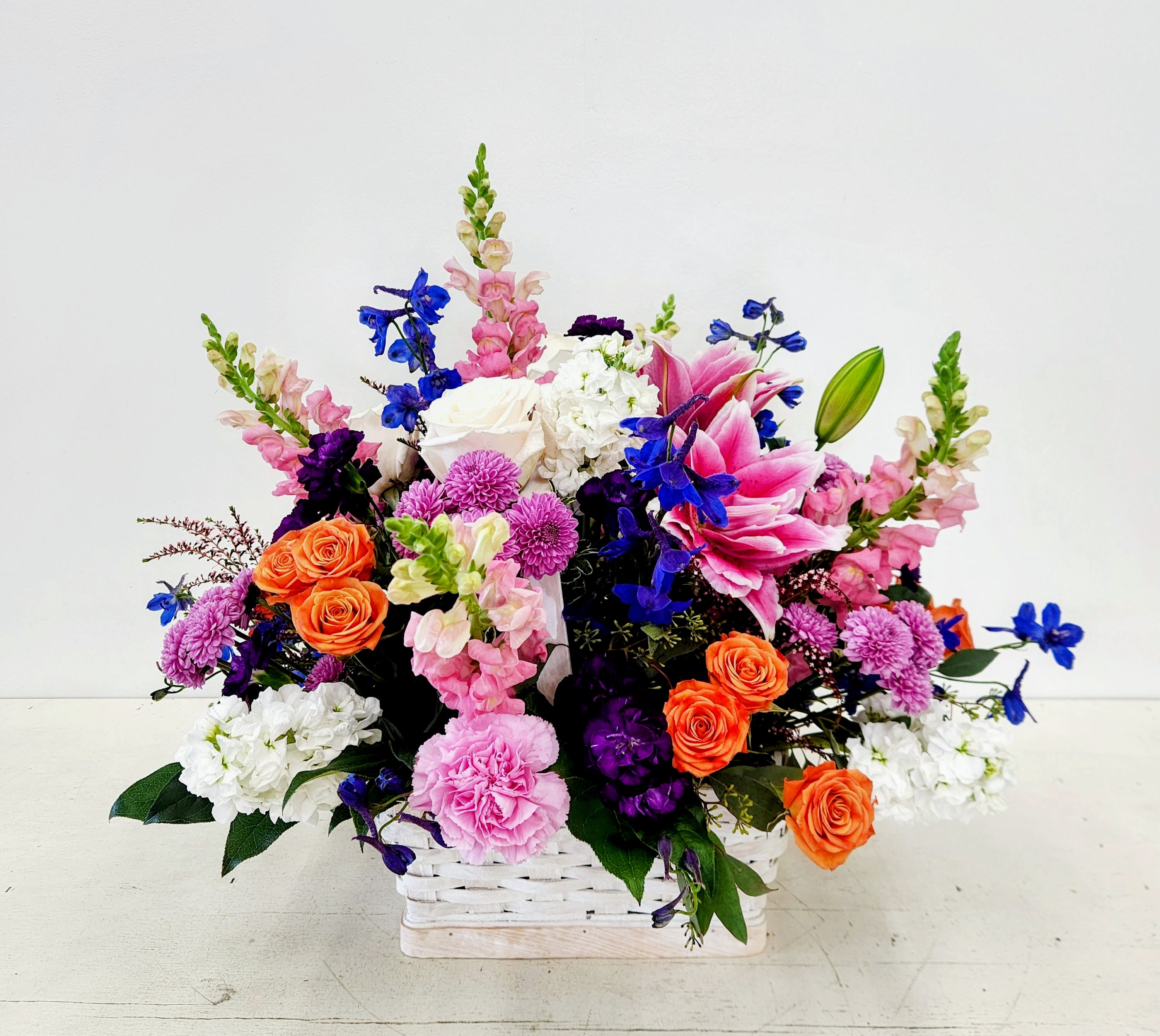 Vibrant Basket of Blooms - A cheerful, color-packed gift that reads like a celebration in bloom - this flower arrangement is a vibrant basket of blooms perfect for a birthday bouquet, congratulations surprise, or a bright thank-you gift. The low, dense design places vivid pink lilies and warm orange roses front and center so the person you're thinking of will feel instantly noticed; the overall palette of pink, purple, orange, white (with touches of blue) feels joyful and uplifting.   