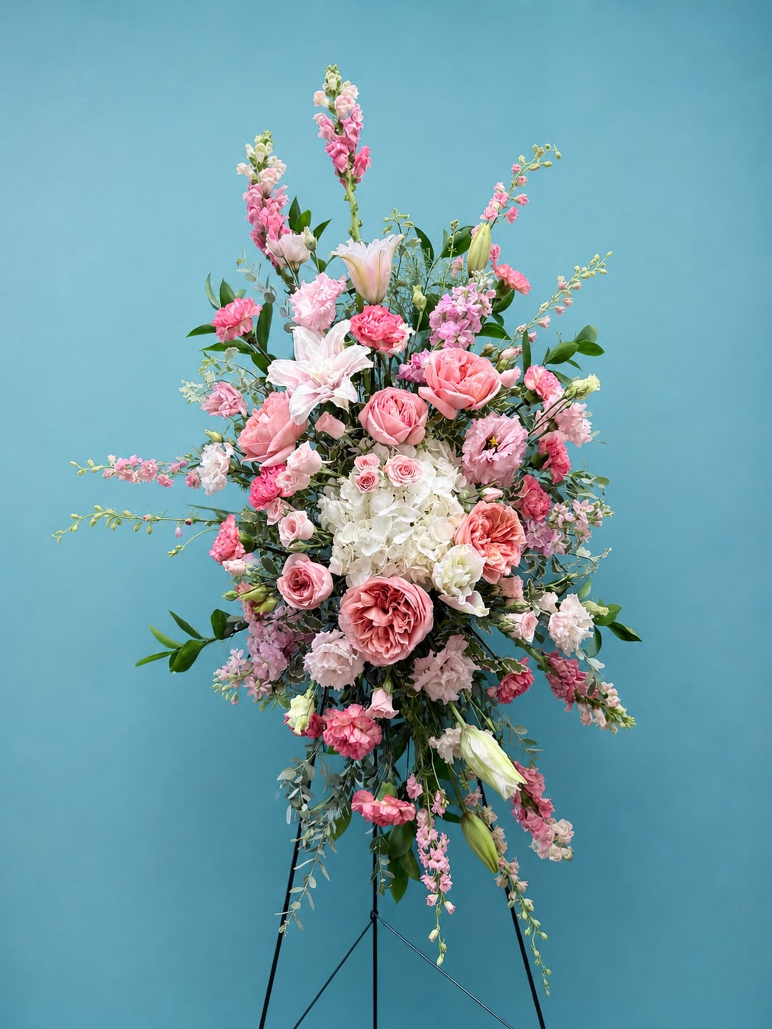 True Sympathy  - A serene standing spray designed for sympathy and funeral services, this oval-shaped arrangement greets the eye with a calm palette of white blooms and soft green accents. The metal easel lifts a layered surface of roses, lilies, hydrangeas and chrysanthemums so the person you're honoring is given a dignified, beautiful focal point that communicates peace and remembrance.  Clusters of white roses and lilies blend with plush hydrangea and textured chrysanthemums, while delicate stock, carnations and small filler flowers add softness. Trailing dendrobium orchid stems and fern fronds extend gently from the composition for a subtle cascading outline; occasional pink and pale-yellow touches bring a whisper of warmth without overwhelming the restful white theme. Displayed on a sturdy black metal easel, the spray presents a full, elongated silhouette appropriate for viewing at a chapel or graveside.  Made to give comfort at memorials, this arrangement conveys sympathy and respect in a refined, timeless way-the kind of tribute that helps the recipient feel seen and supported. Available in a tall easel format for prominent display; standard care is minimal once arranged, and our florists will ensure the spray is ready for delivery or funeral home pickup.