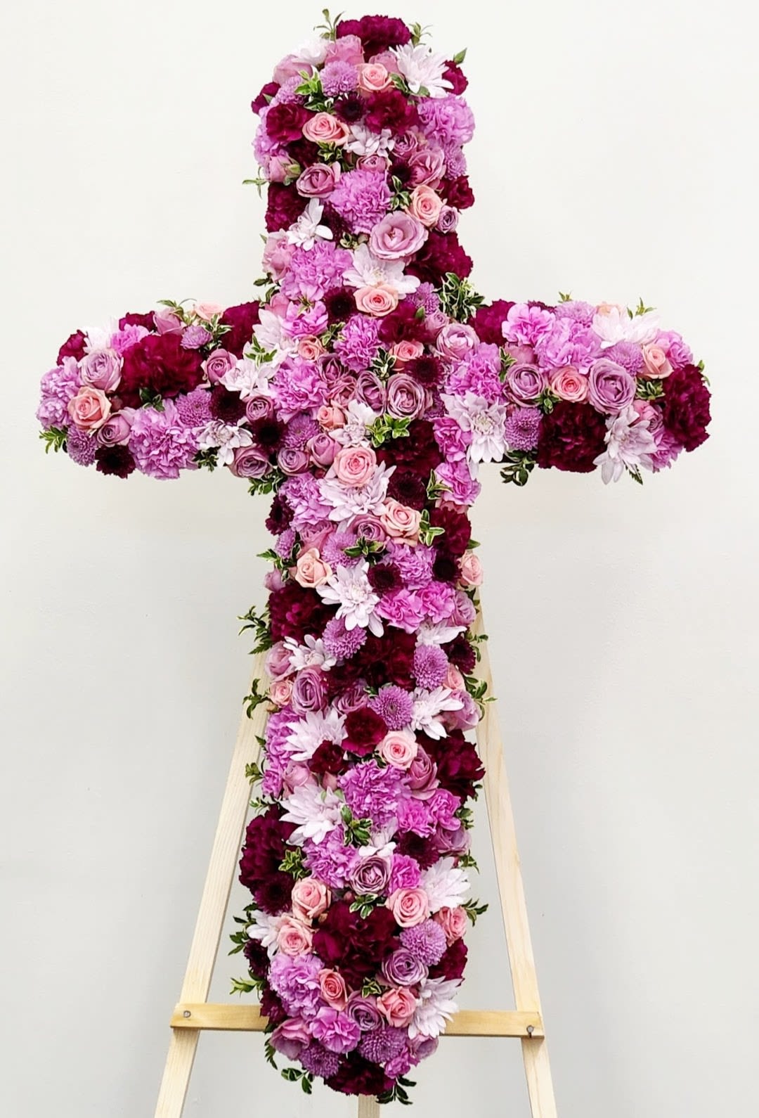Purple and Lavender Cross - A large, dignified floral cross in soft pinks, lavender, whites and deep burgundy creates a reverent tribute for a funeral or memorial service. This sympathy arrangement is designed as a standing tribute on a wooden easel so the person you're honoring will be presented with a full, ceremonial display that reads as both comforting and respectful. The balanced, symmetrical shape and layered color play convey a gentle solemnity that helps express sympathy and remembrance.  