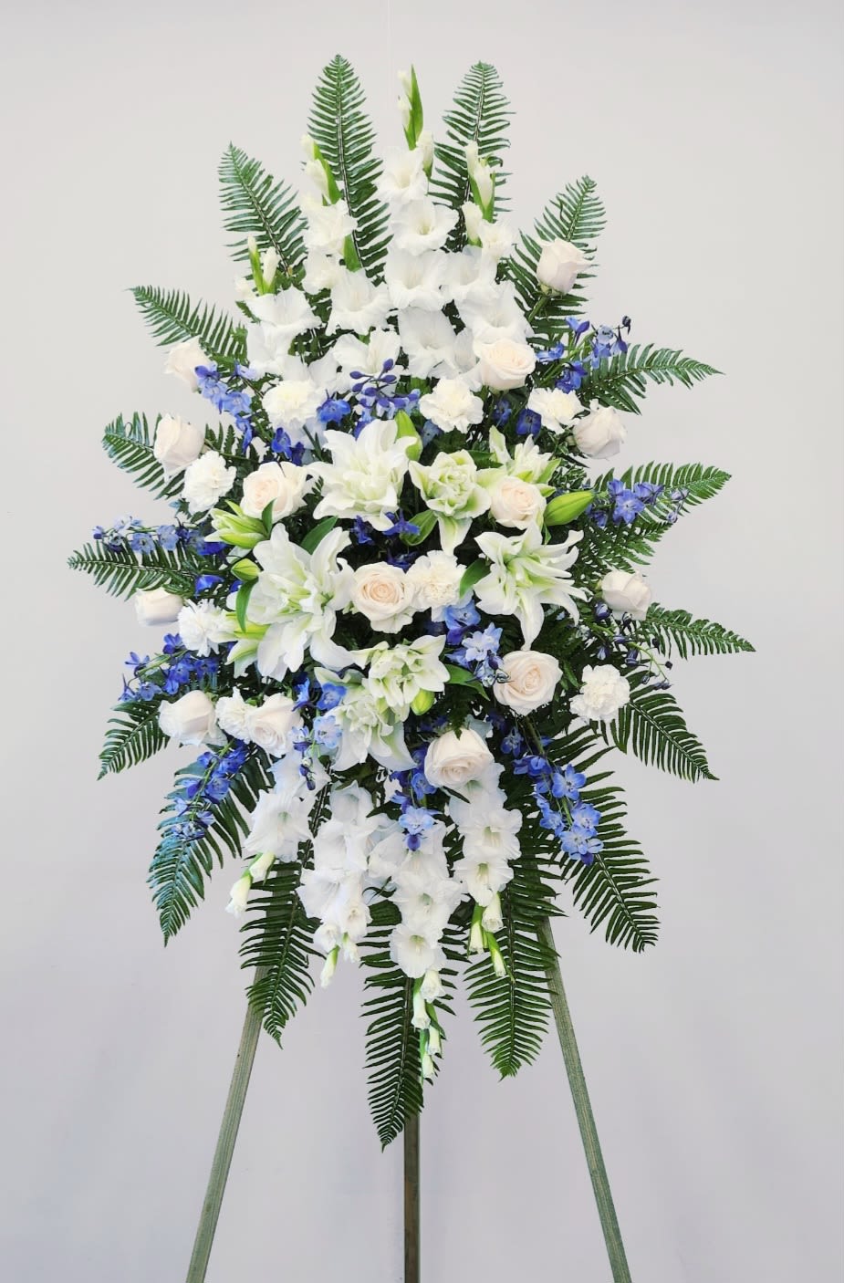 Ocean Song Spray - A tall, dignified standing spray in cool white, blue, and green tones that offers a serene tribute for a funeral or memorial service. This sympathy arrangement features white lilies, roses, and carnations layered with sprays of blue delphiniums and lush fern fronds to create a formal, symmetrical silhouette that reads respectful and comforting from front pew to back row.  