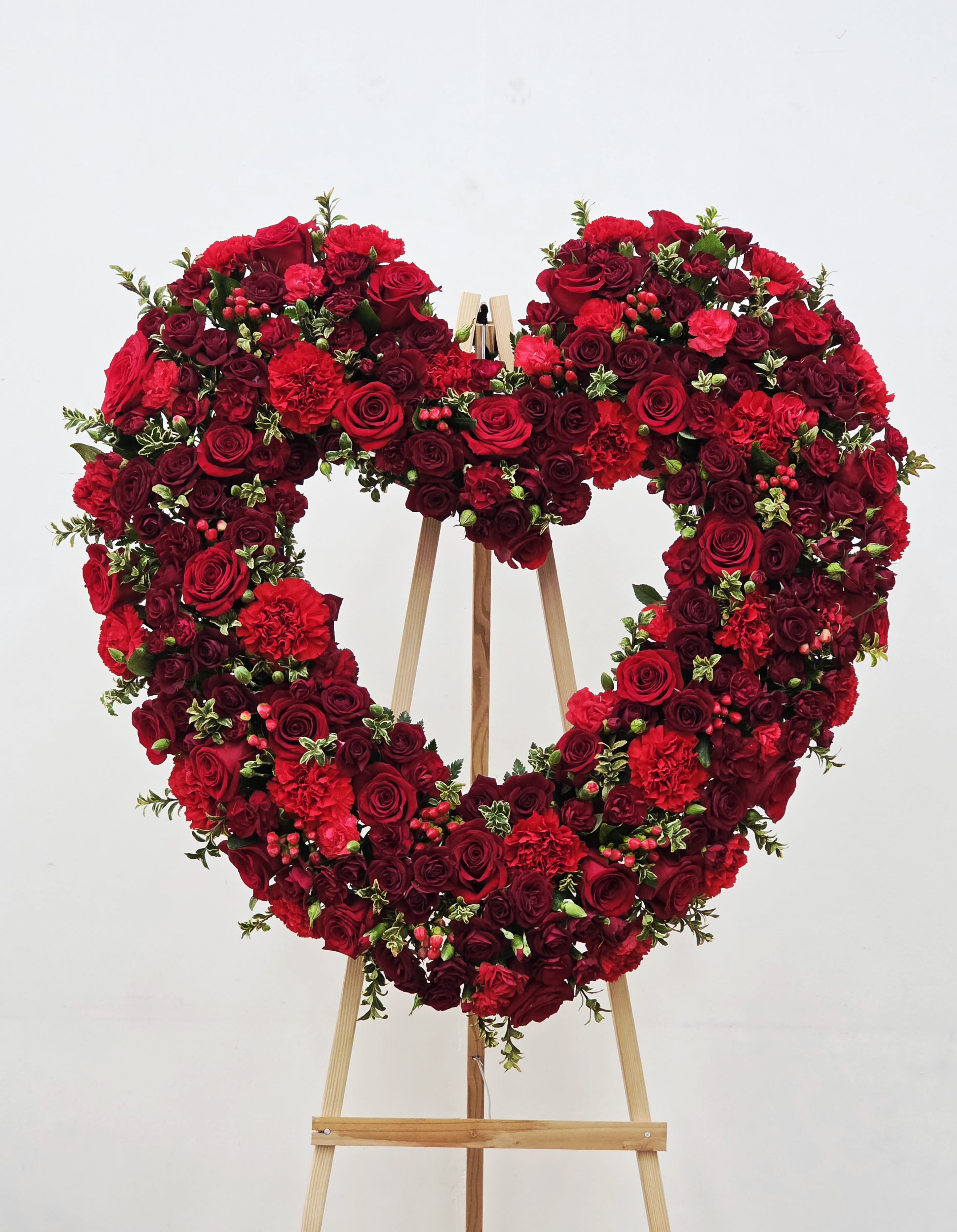 Love and Devotion Heart Spray - A dense heart of red and burgundy roses, accented with bright red carnations, creates an unmistakable statement of love or remembrance in this standing heart wreath. Designed as a large heart-shaped arrangement on a wooden easel, it reads ceremonial-an ideal choice for an anniversary tribute, or a respectful memorial display for the person you're honoring.  
