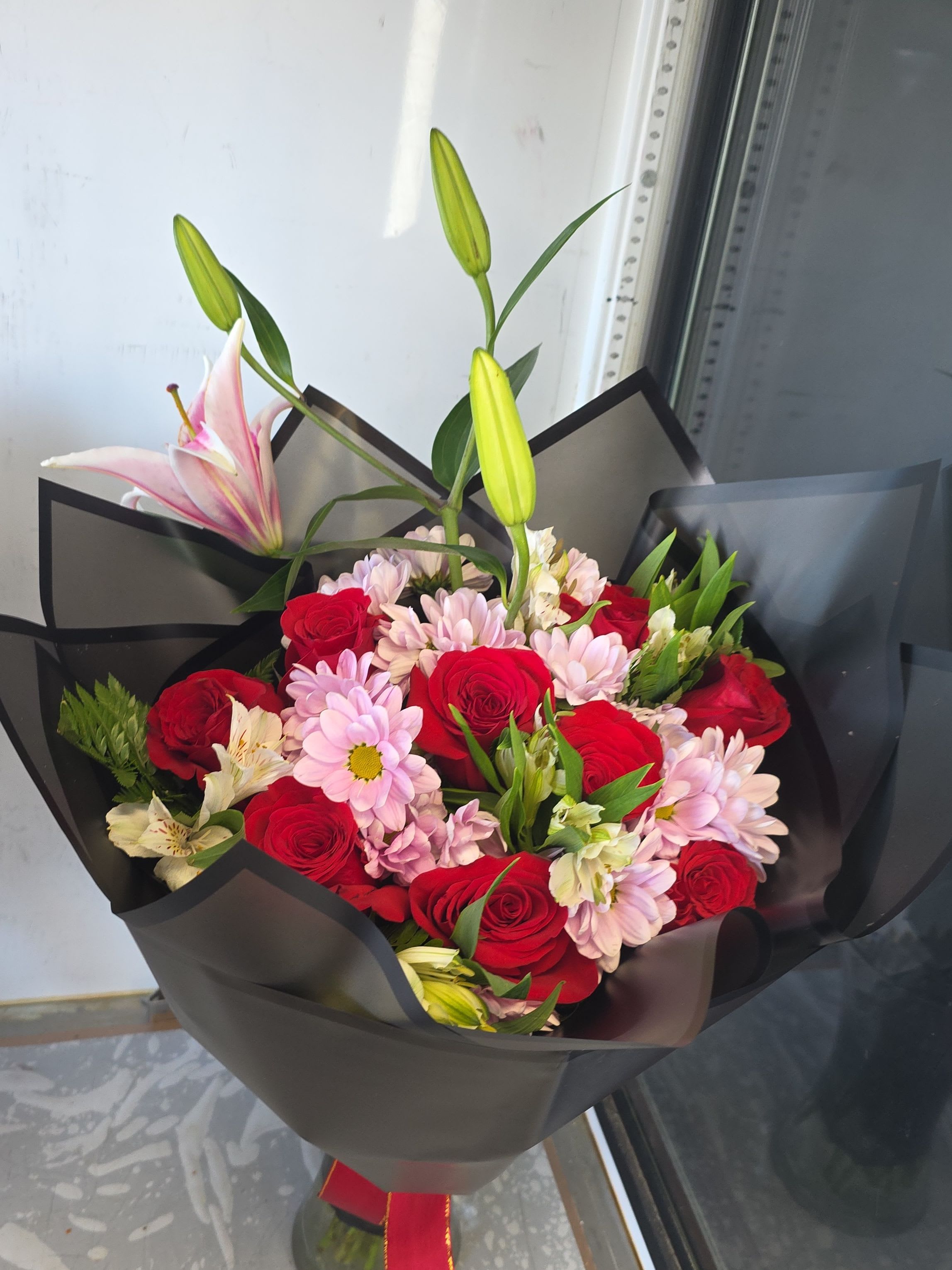 Bouqueth rosas rojas mixto  - A compact cluster of deep red roses and pale pink chrysanthemums makes a bold, romantic statement in this red-and-pink bouquet-perfect as an anniversary bouquet, Valentine's Day gift, or a "thinking of you" surprise. White alstroemeria threads through the arrangement for delicate contrast, while a few tall pink lily stems and unopened buds rise above the cluster to add graceful height and the promise of unfolding fragrance.  Wrapped in sleek black bouquet paper and tied with a red ribbon, the modern styling sharpens the color contrasts and gives the arrangement a polished, contemporary look ideal for Mother's Day, birthdays, graduations, or to bring comfort at sympathy and funeral services. The mix of roses, chrysanthemums, alstroemeria, and lilies balances lush texture and long-lasting bloom life; choose this for romantic gestures, housewarmings, get-well wishes, or to make someone feel truly seen.  This design ships as a hand-tied bouquet-compact enough to hold but generous in visual impact. To prolong bloom life, re-cut stems and place in fresh water upon arrival; lilies will open over several days, extending the bouquet's life and fragrance.