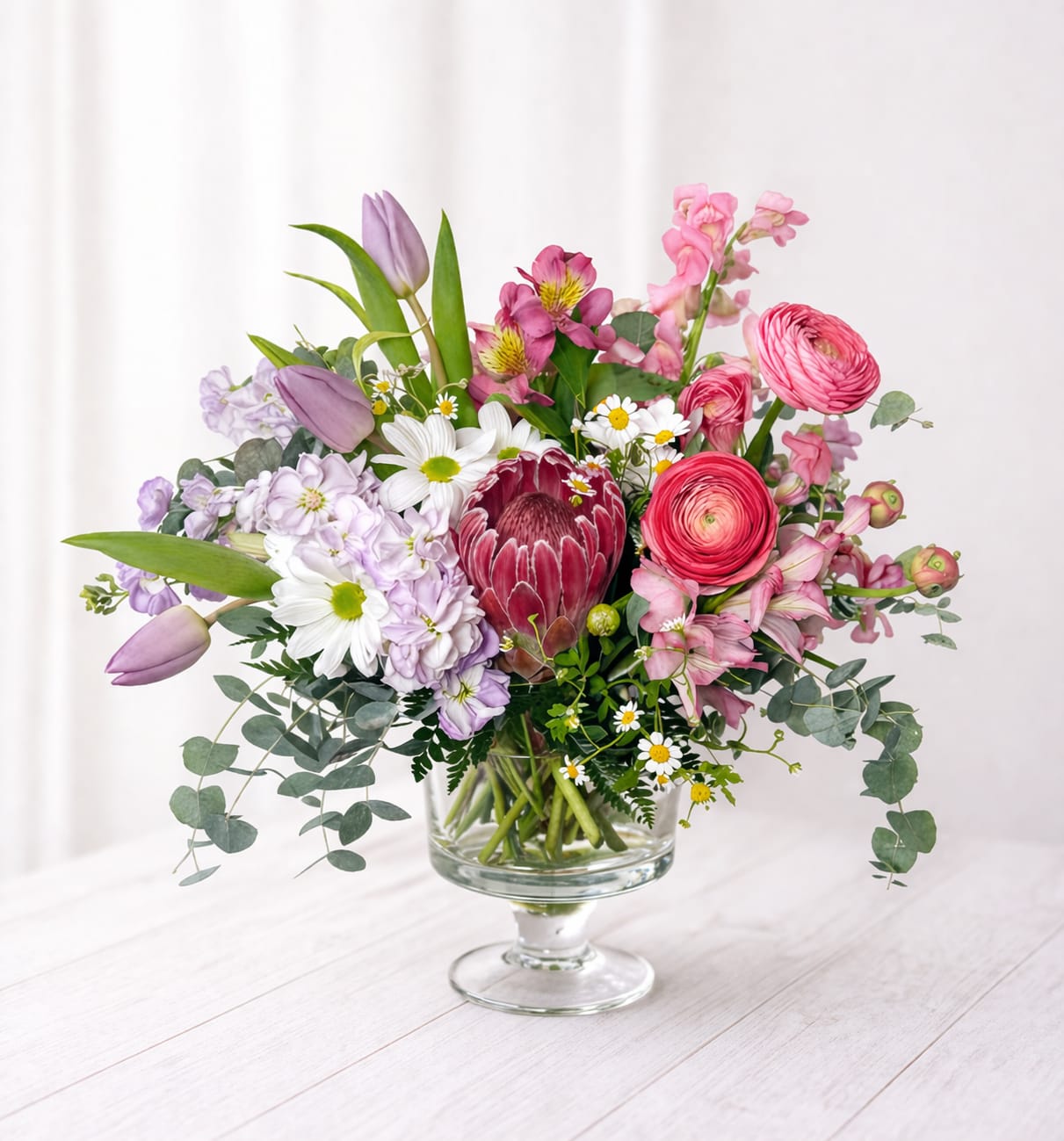 Meadow Garden Vase - Blushing Meadow features a romantic mix of pink ranunculus, lavender tulips, delicate daisies, lush hydrangea, and a striking protea focal—balanced with airy greenery for that effortless, freshly gathered look. Designed in a clear glass vase, this piece is perfect for birthdays, celebrations, or simply brightening someone’s day with something truly special.