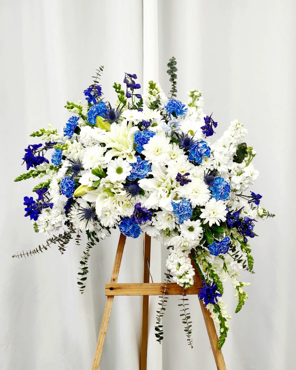 Blue and White Crescent Spray - A sweeping crescent of blue and white blooms gives this standing spray a serene, ceremonial presence-perfect as a sympathy arrangement or memorial tribute to honor a life. The design reads formal and uplifting: a dense, rounded center of white lilies and carnations flows into cascading side elements that gently curve, so the person you're remembering will be given a composition that feels both respectful and full of grace. 