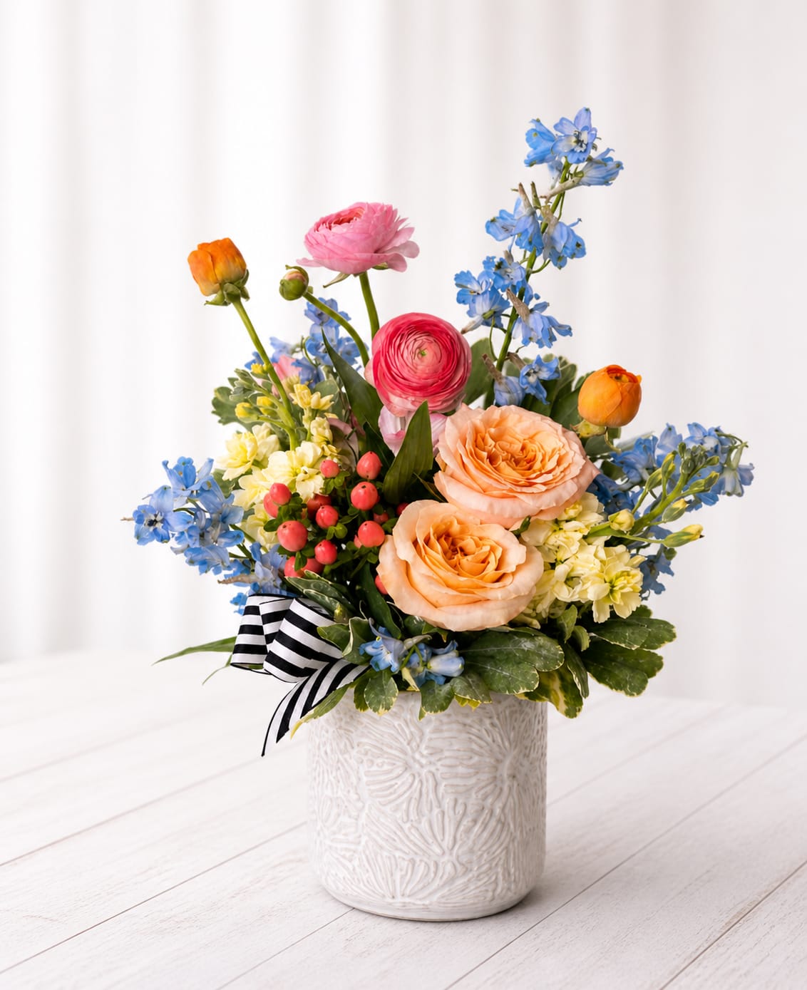 Sunrise Garden - Bright, cheerful, and full of personality—Sunrise Garden is a playful mix of soft peach roses, vibrant ranunculus, delicate blue delphinium, and pops of seasonal blooms, all gathered into a textured ceramic keepsake container. The fresh color palette of peach, coral, and sky blue brings a happy, uplifting feel—perfect for celebrating life’s little (and big) moments. Finished with a classic black-and-white ribbon for a touch of charm, this arrangement is ideal for birthdays, thank-yous, or just because.