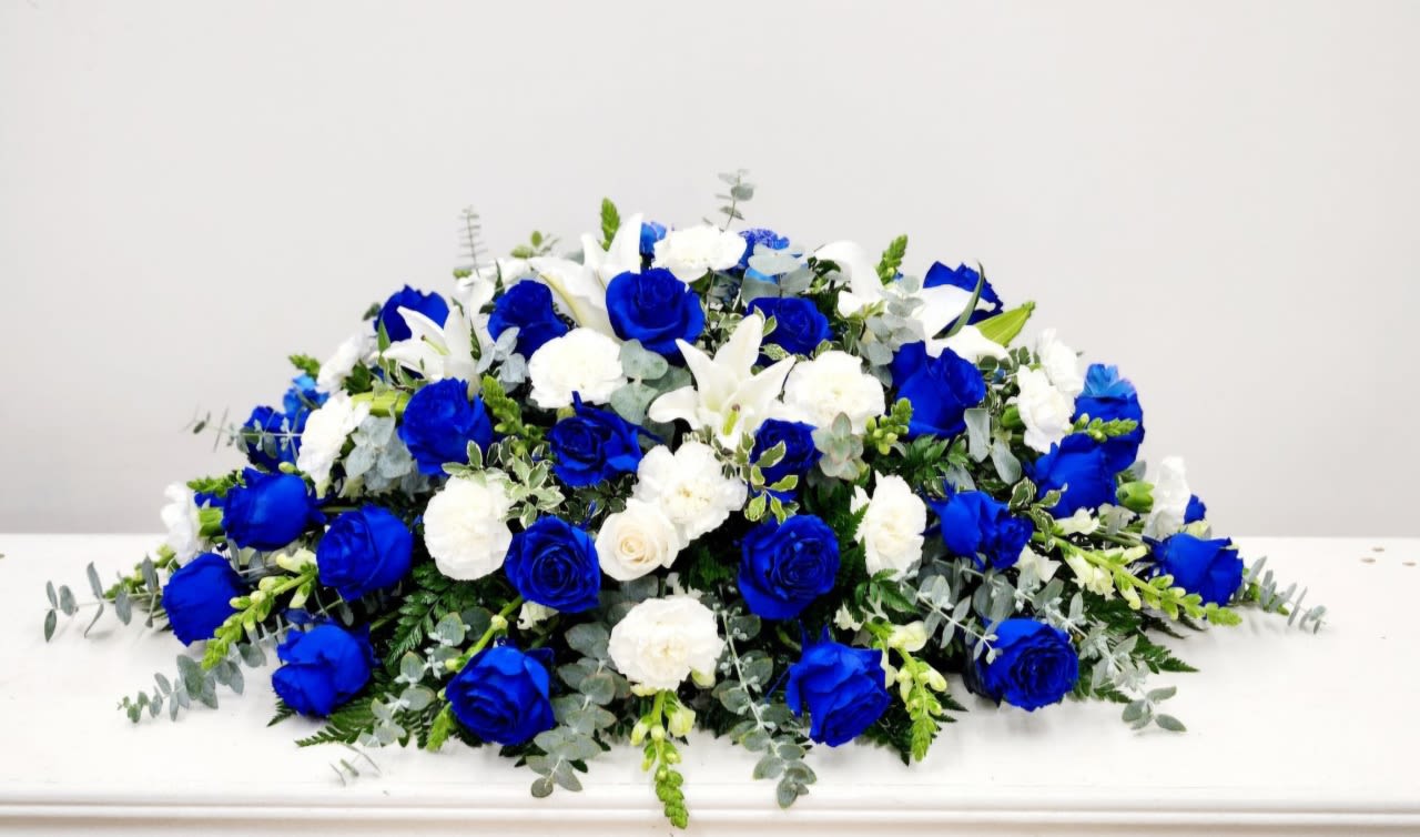 Cherished and Remembered  Casket Spray - Royal-blue and white blooms form a serene, dignified spread perfect for a sympathy or memorial casket spray. The cool blue-and-white palette-vivid royal-blue roses paired with crisp white roses and open white lilies-creates an immediate visual impression that feels both respectful and heartfelt, the kind of arrangement that helps the person you're honoring feel deeply remembered.  
