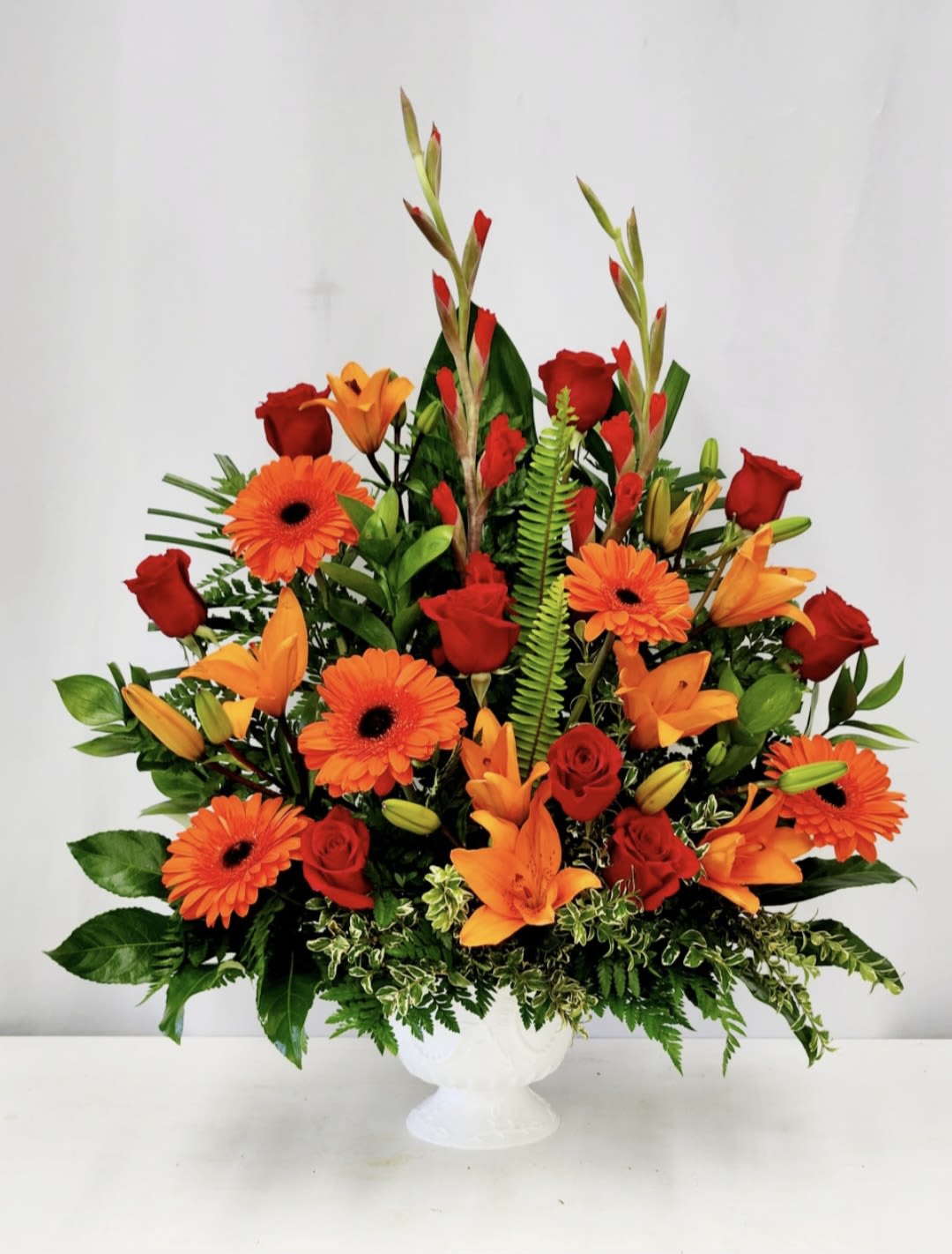 Vibrant Memory - Bright red roses, orange gerbera and lilies arranged full and upright in a white ceramic pedestal vase. PREMIUM SIZE ONLY. 25" by 32"  