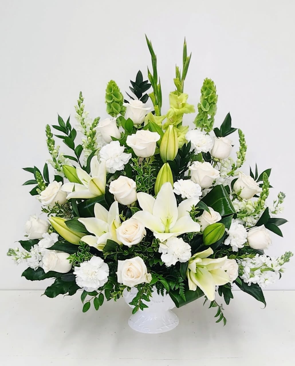 White and Green Fan Shaped Arrangement - Large symmetrical fan of white lilies, roses, carnations and green snapdragons in a white pedestal vase, accented with beautiful greens that brings out the beauty of the white blooms. 