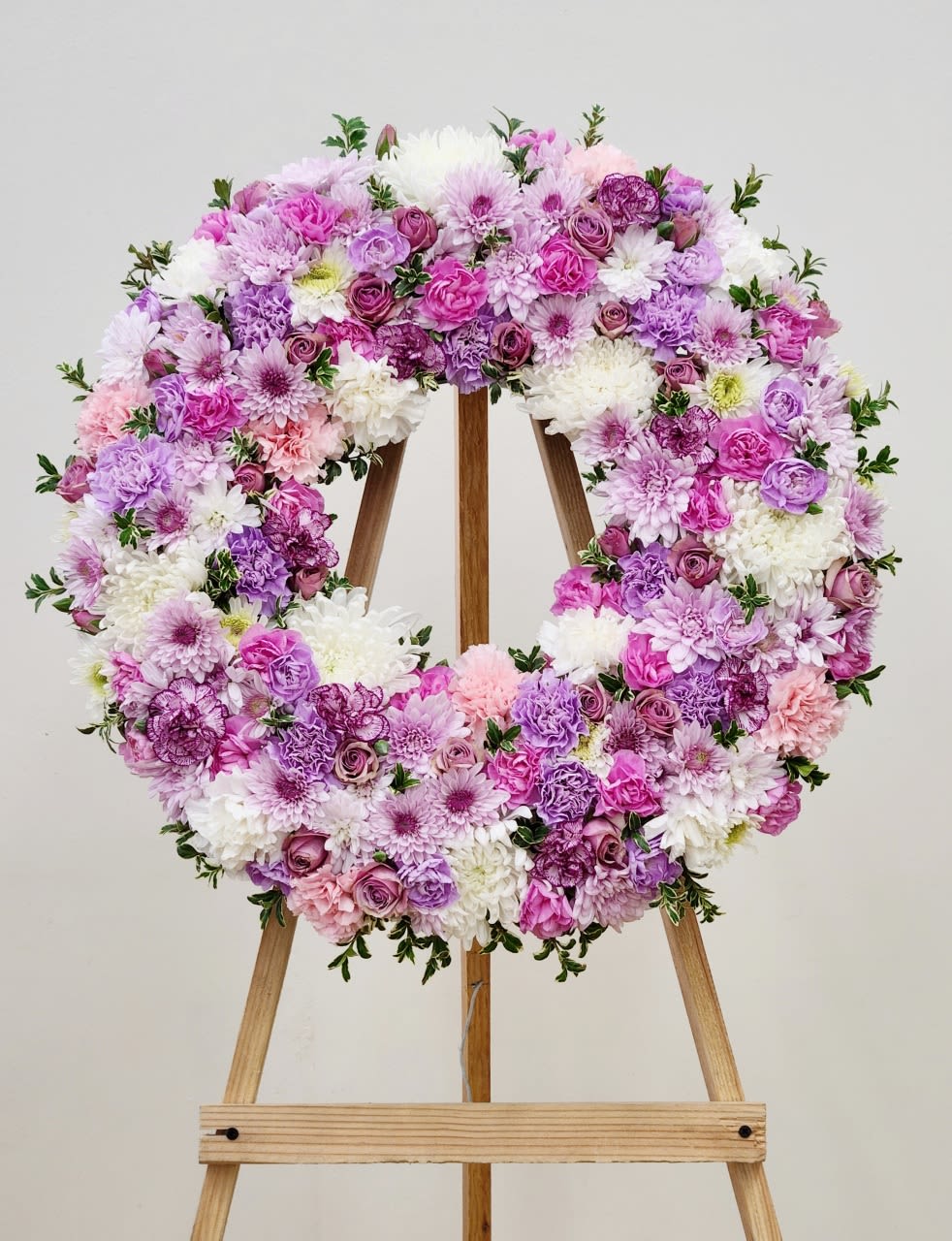 White and Lavender Wreath - Large circular wreath of mixed pink, lavender, purple, and white blooms on a wooden easel, full and textured with soft greens all around the wreath.