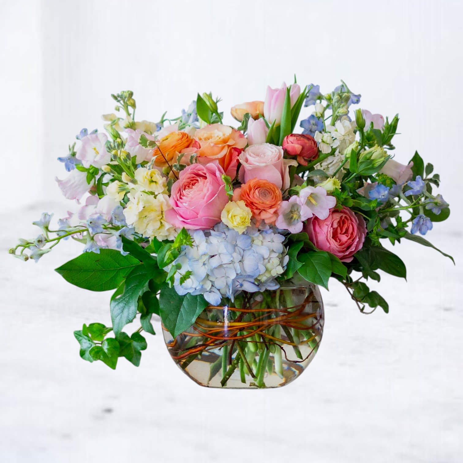 Pastel Poetry - Soft, airy, and overflowing with garden-inspired beauty, Pastel Poetry features a dreamy blend of blush and peach roses, delicate ranunculus, airy delphinium, hydrangea, tulips, and whimsical seasonal blooms. Designed with natural movement and layered textures, this arrangement feels romantic, artistic, and effortlessly elegant.  Arranged in a clear glass vase wrapped with natural vine accents, this design is perfect for birthdays, anniversaries, baby showers, sympathy gestures, thank-you gifts, or simply celebrating everyday beauty.  Available for same-day flower delivery in Galveston, Jamaica Beach, Tiki Island, Sea Isle, Hitchcock, La Marque, Texas City, Dickinson, League City, Kemah, Clear Lake, Webster, and surrounding coastal areas.