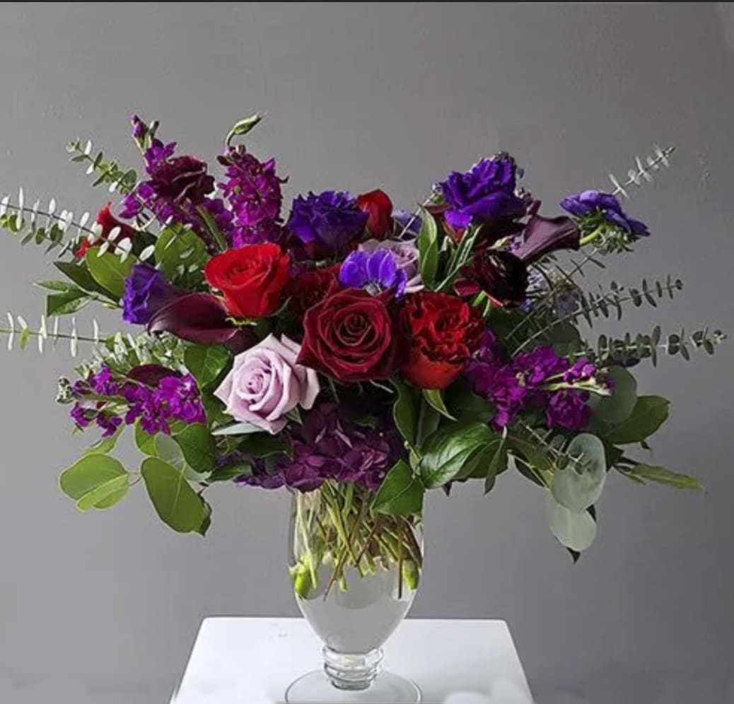 A Victorian Dream (PREORDER ONLY) - Product Description: Capturing the beauty of the royal flower beds in Victorian England, this Exquisite design glows with jewel tone hues and rich texture. Red heart roses, black bacarra roses, ocean song roses, red piano garden roses, purple mini calla lilies,  purple lisianthus, on a bed of fluffy purple hydrangea. Adorned with baby blue eucalyptus and parvi eucalyptus. Arranged in a tall tapered footed compote.