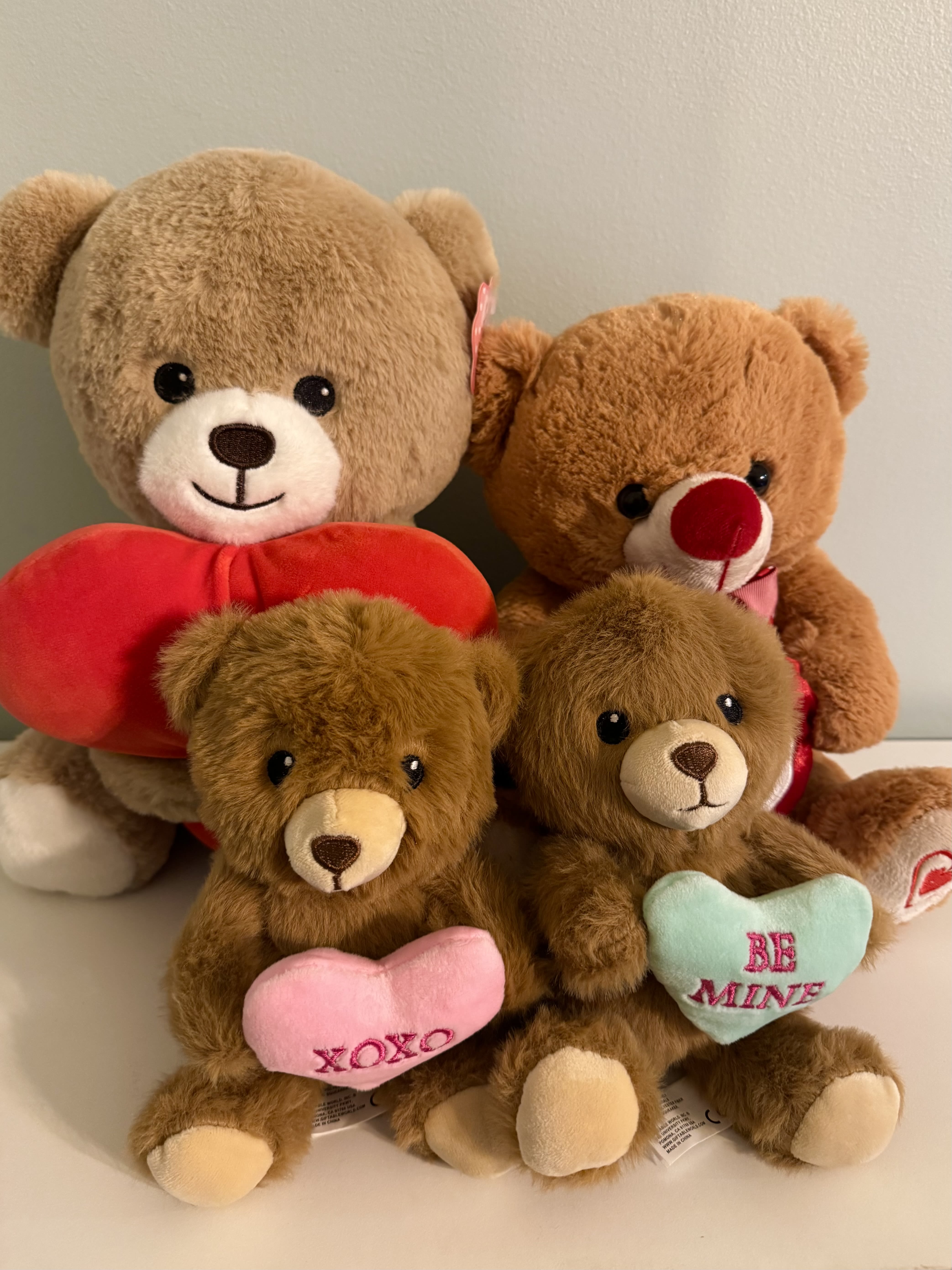 "Mother`s Day" Special Item #3 - A cluster of cuddly plush teddy bears makes this flower arrangement an affectionate choice for Mother's Day or a romantic gift for Valentine's Day-perfect when you want the person you're thinking of to feel warm, cherished, and playfully adored. This gift-forward arrangement reads like a soft embrace: four close-set bears in shades from tan to deeper brown, each clutching a heart-shaped plush pillow embroidered with messages like "XOXO" and "BE MINE," so the recipient sees your sentiment at once.  Rather than traditional stems, the focus here is texture and color-warm browns, pinks, reds and a touch of blue combine to create a cozy, cheerful palette that works well for moms who treasure keepsakes or anyone who loves whimsical, plush-centered gifts. The bears are arranged compactly against a light background for a clean, gift-box look; the heart pillows introduce a romantic accent without feeling overstated.  Choose this piece when you want to mark Mother's Day, an anniversary, or a heartfelt "thinking of you" moment with something that lasts. It's ideal for a bedside table or living-room display where the recipient will keep returning to that tactile reminder of your care. Order with standard delivery timing and no special care is required beyond keeping the plush clean and dry.