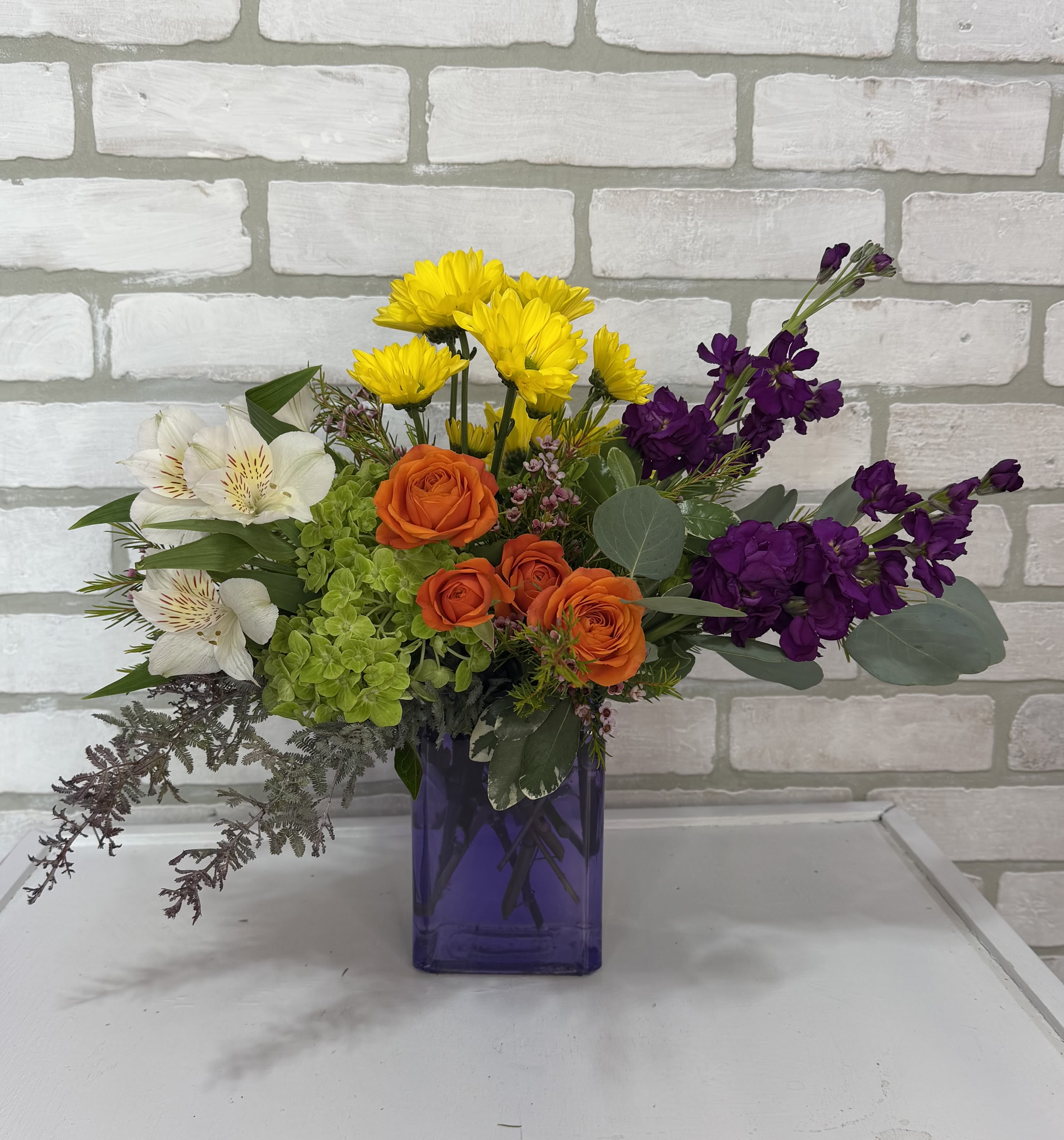 Mama Mia - A purple vase arranged with daisies, alstroemeria lilies, stock, green hydrangea, and spray roses.