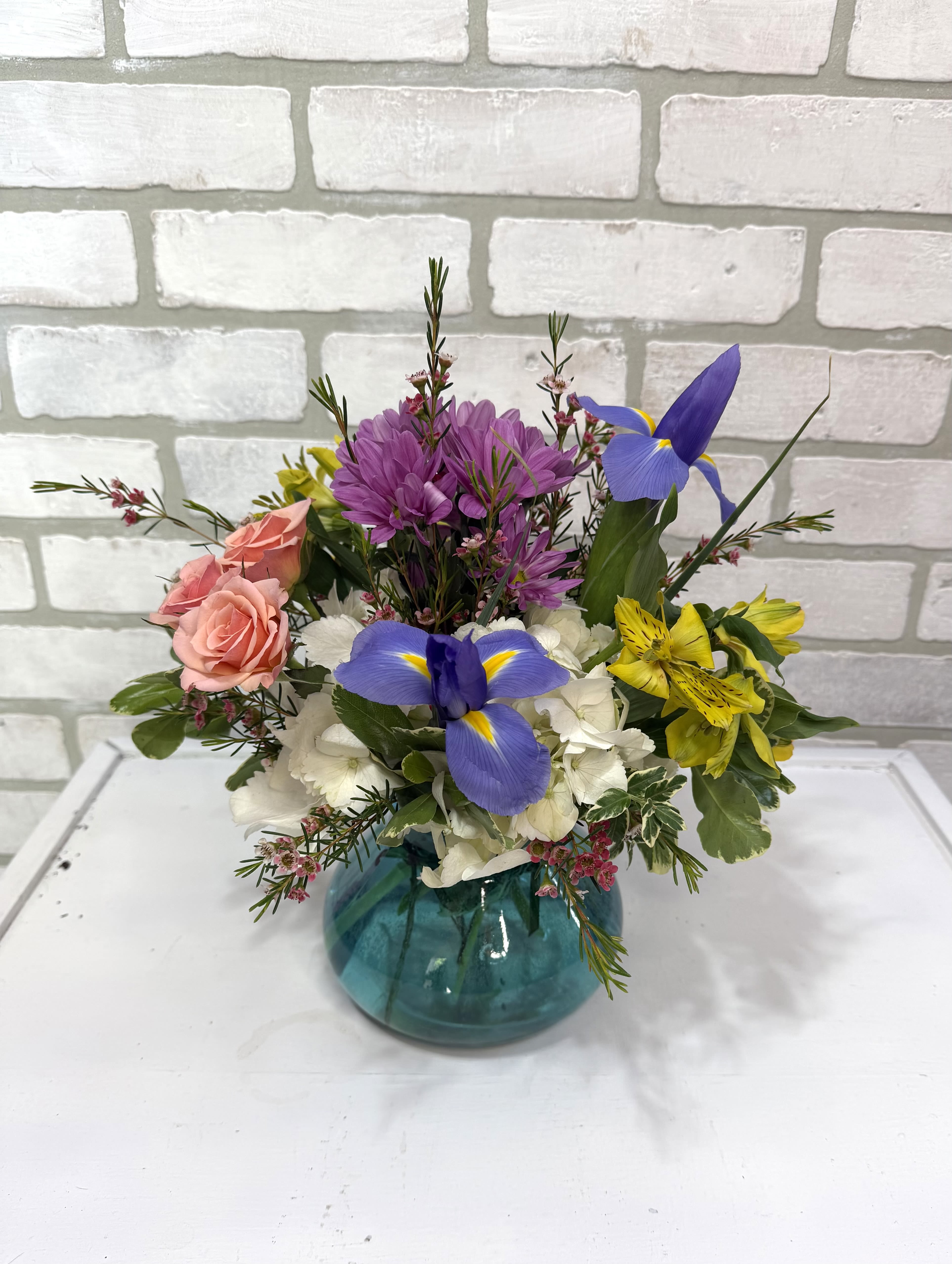 Mixed bouquet in a teal glass vase with pink roses, purple blooms, and blue iris