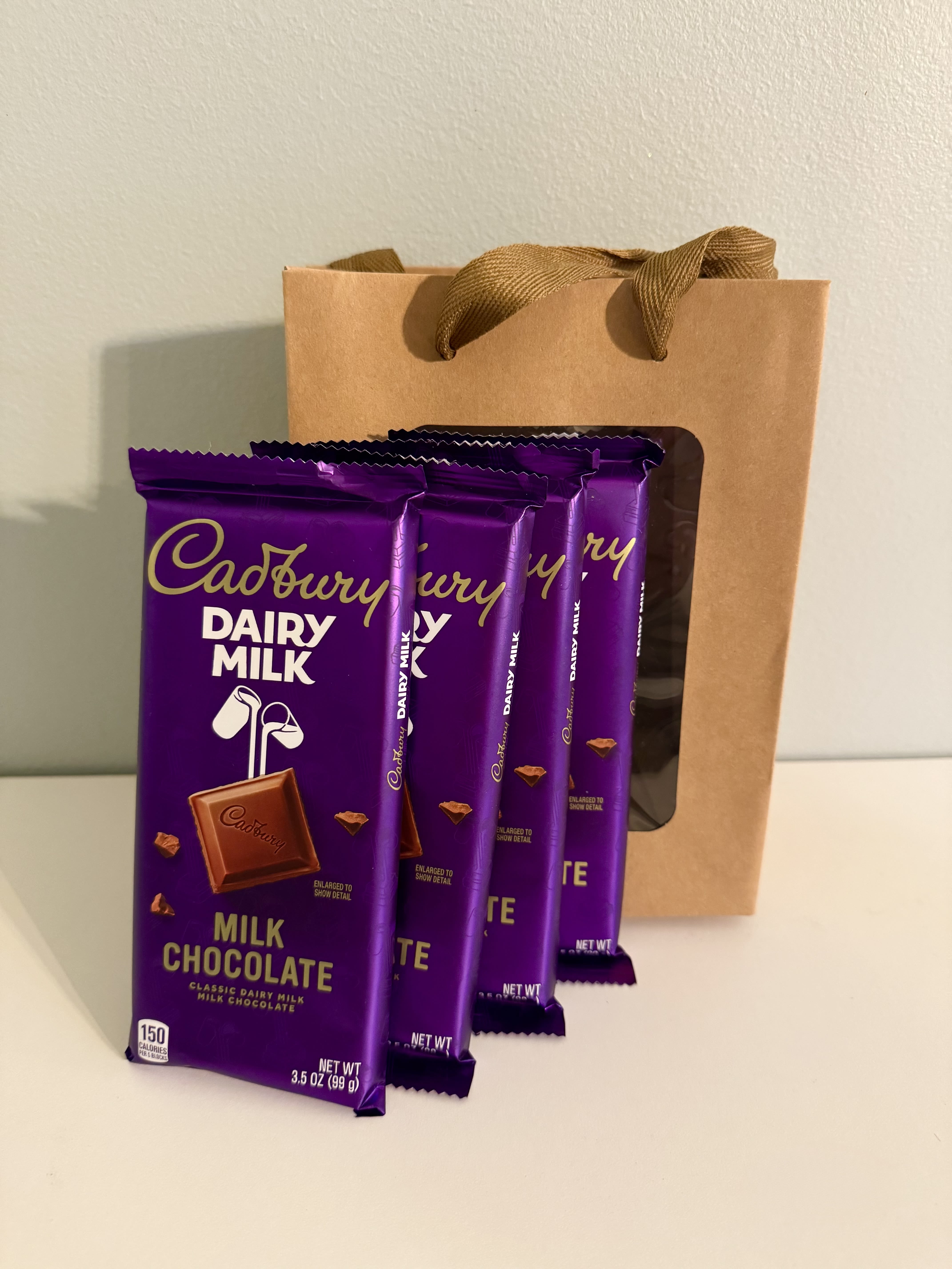 "Mother`s Day" Special Item #6 - A bold pop of Cadbury purple makes this Mother's Day gift set feel joyful and indulgent - perfect as a small Mother's Day treat or add-on for a birthday or thank-you. As a flower arrangement companion or stand-alone gift, it sends a clear message: you're thinking of her and want to brighten her day with something sweet and celebratory.  This set includes several Cadbury Dairy Milk chocolate bars neatly presented in a kraft paper gift bag with ribbon handles. The deep purple wrappers contrast beautifully with the warm tan bag for an elegant, modern look that pairs well with lavender or purple-toned blooms. The presentation is simple and gift-ready, sized for easy hand delivery or to tuck alongside a bouquet.  Give this to a chocolate-loving mom, sister, teacher, or friend who deserves a small indulgence - the sort of treat that makes someone feel seen and appreciated. Order as an add-on to any bouquet or as a standalone sweet surprise; no special care needed beyond keeping the chocolates cool and dry until gifting.