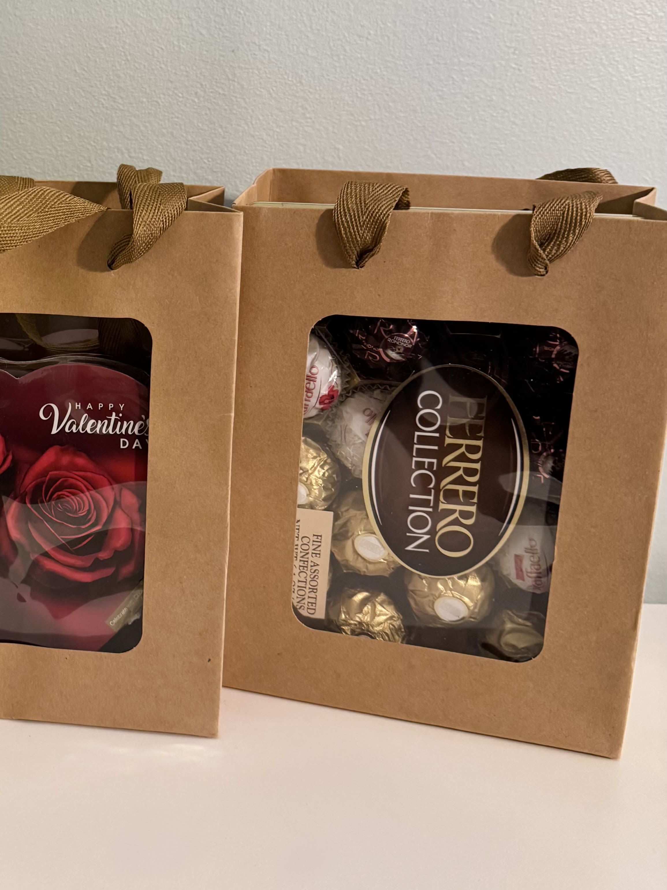 "Mother`s Day" Special Item#8 - Two kraft paper gift bags packed with assorted chocolates and candy make this Mother's Day gift feel warm and personal-perfect as a small Mother's Day gift, anniversary treat, or a thoughtful thank-you surprise. The clear window panels and ribbon handles show off rich, dark confections (including a Ferrero Collection box and a Valentine-themed pack with a red rose graphic), so the person you're celebrating will see the indulgence before they even open it. The brown, red, black, and gold palette reads romantic and classic, so it works just as well for a cozy Valentine's moment or a simple "thinking of you" sweet.  Each bag arrives ready to give: kraft paper with ribbon handles and a neat, gift-ready presentation that highlights the chocolates inside. Because these are pre-packed sweet-goodie bags rather than a floral arrangement, they're an easy add-on for anyone who wants to pair treats with flowers or send a standalone edible surprise. Order as a pair or add to a bouquet to create a fuller Mother's Day or anniversary package-no special care required beyond keeping the sweets in a cool, dry place until gifting.