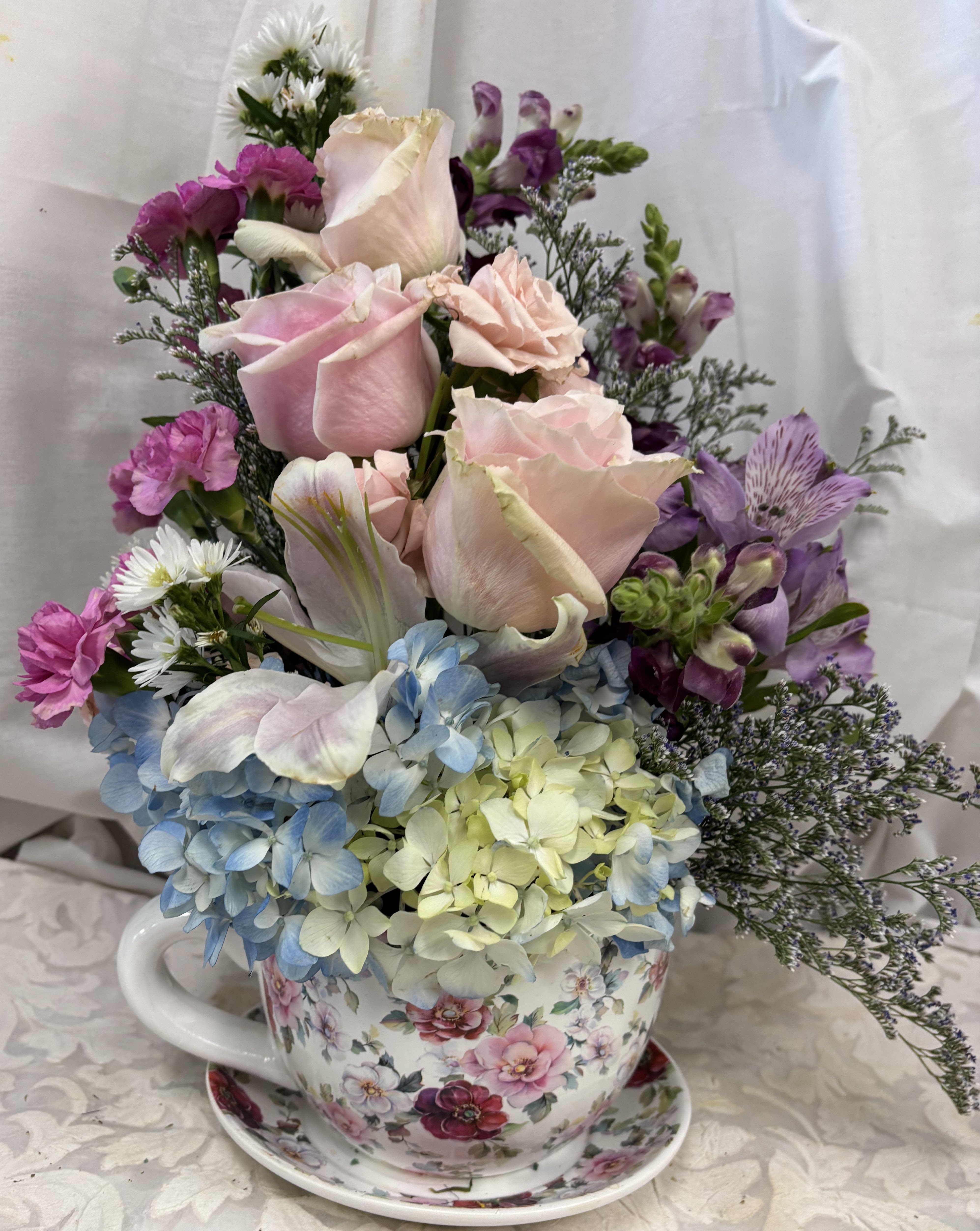 Moms Rule - In a planter sized teacup all the strong garden flowers fill this pretty teacup.  Roses, spray carnations, hydrangeas, liles, alstromerias fill spill over this cup along with seasonal greenery.