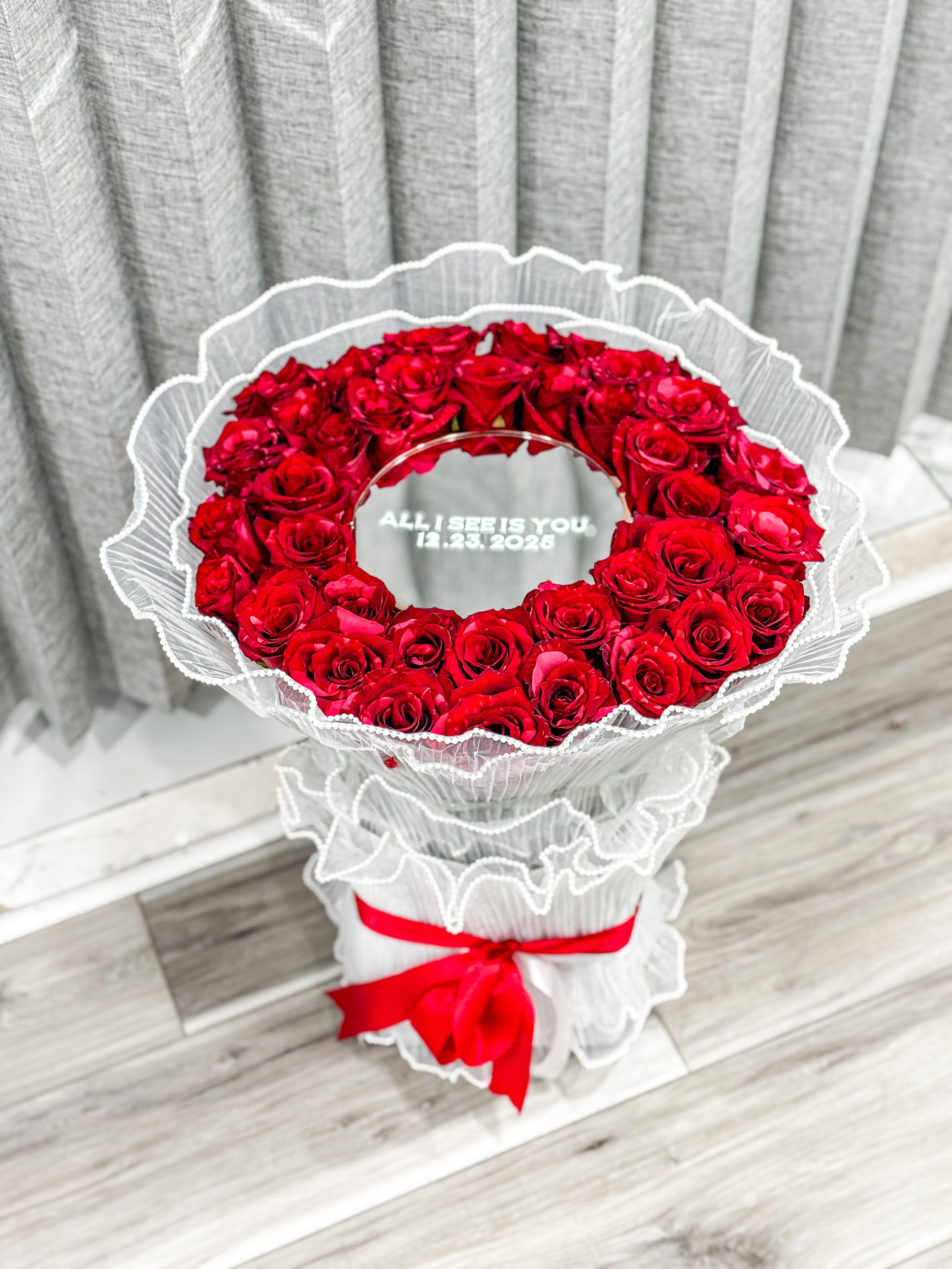 Forever Reflection- Mirror Rose Bouquet - A dramatic ring of deep red roses arranged in a perfect circular wreath-this anniversary bouquet and romantic gift makes the person you're thinking of feel truly honored. The Forever Reflection-Mirror Rose Bouquet reads like a keepsake: tightly packed garden roses form a polished, dome-like ring with an open center that can hold a clear acrylic sign or a printed date for commemorative moments, so it's equally right for anniversaries, Valentine's Day, weddings, or a meaningful Mother's Day tribute.  Every stem is wrapped in layered white mesh or tulle and finished with a bright red satin ribbon for a formal, high-impact presentation that emphasizes the vivid red petals and clean white wrap. The compact, structured shape gives a luxe, tailored look ideal for sympathy and memorial displays as well as celebratory events like graduations, birthdays, or housewarmings. Available in sizes suited to intimate gifts or larger statement pieces; delicate care-fresh water and a cool spot away from direct sun-will keep the roses looking their best for special-day moments and beyond.
