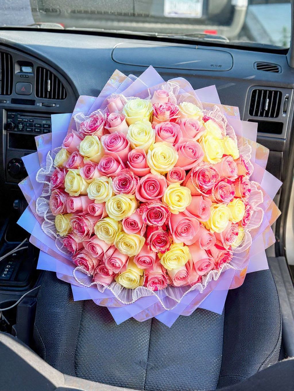 Eternal Love Ring- Signature Bouquet - A lush, tightly packed hand-tied bouquet of roses in soft pinks, pale yellow, cream and white-perfect as a birthday bouquet, anniversary roses gift, or a radiant Mother's Day surprise. The Eternal Love Ring- Signature Bouquet reads as a substantial, romantic presentation: the rounded mass of blooms creates an immediate impression of abundance and tenderness that will make the person you're thinking of feel genuinely cherished.  Each bloom is gathered close for a dense, luxurious look and wrapped in layered lavender and pastel paper with sheer decorative mesh and a ruffled trim for an ornate, gift-ready finish. Tulle ribbon and clear decorative mesh add delicate texture and keep the bouquet feeling airy and feminine while the color palette stays bright and celebratory. Shown large and hand-tied, this arrangement arrives ready to carry and present-no vase needed-making it an easy, elegant choice for anniversaries, Valentine's Day, birthdays, or any moment you want to mark with warmth and joy.