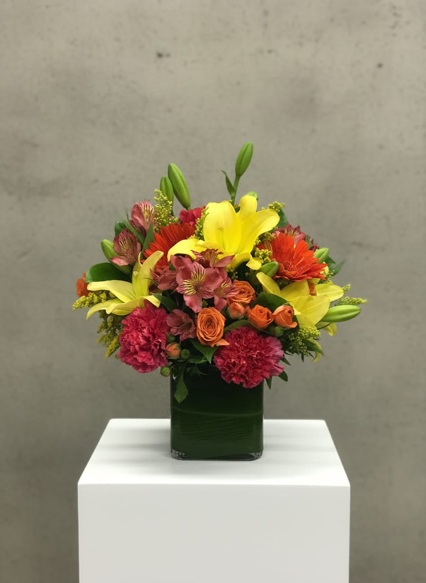 Sunburst Symphony - “Sunburst Symphony” feels like a celebration captured in a vase—bright, lively, and impossible to ignore. The arrangement bursts with color, anchored by radiant yellow lilies that open like little suns, their petals stretching outward and setting the tone for the whole design. Around them, clusters of orange spray roses add warmth and texture, their smaller blooms creating a soft, glowing contrast.  Hot pink carnations and alstroemeria weave through the arrangement, bringing bold pops of color that feel playful and energetic. The carnations give a ruffled, full-bodied look, while the alstroemeria add delicate, star-like accents that keep the eye moving. Sprigs of solidaster are tucked throughout, offering airy touches of golden yellow that lighten the composition and add a hint of wild, natural charm.  Bright orange gerbera daisies stand out like cheerful faces, their wide, open blooms adding a modern, graphic element that ties the whole palette together. Everything is gathered into a clean, cubed glass vase, where a neatly arranged green leaf lining wraps the inside, hiding the stems and giving the design a polished, fresh finish.