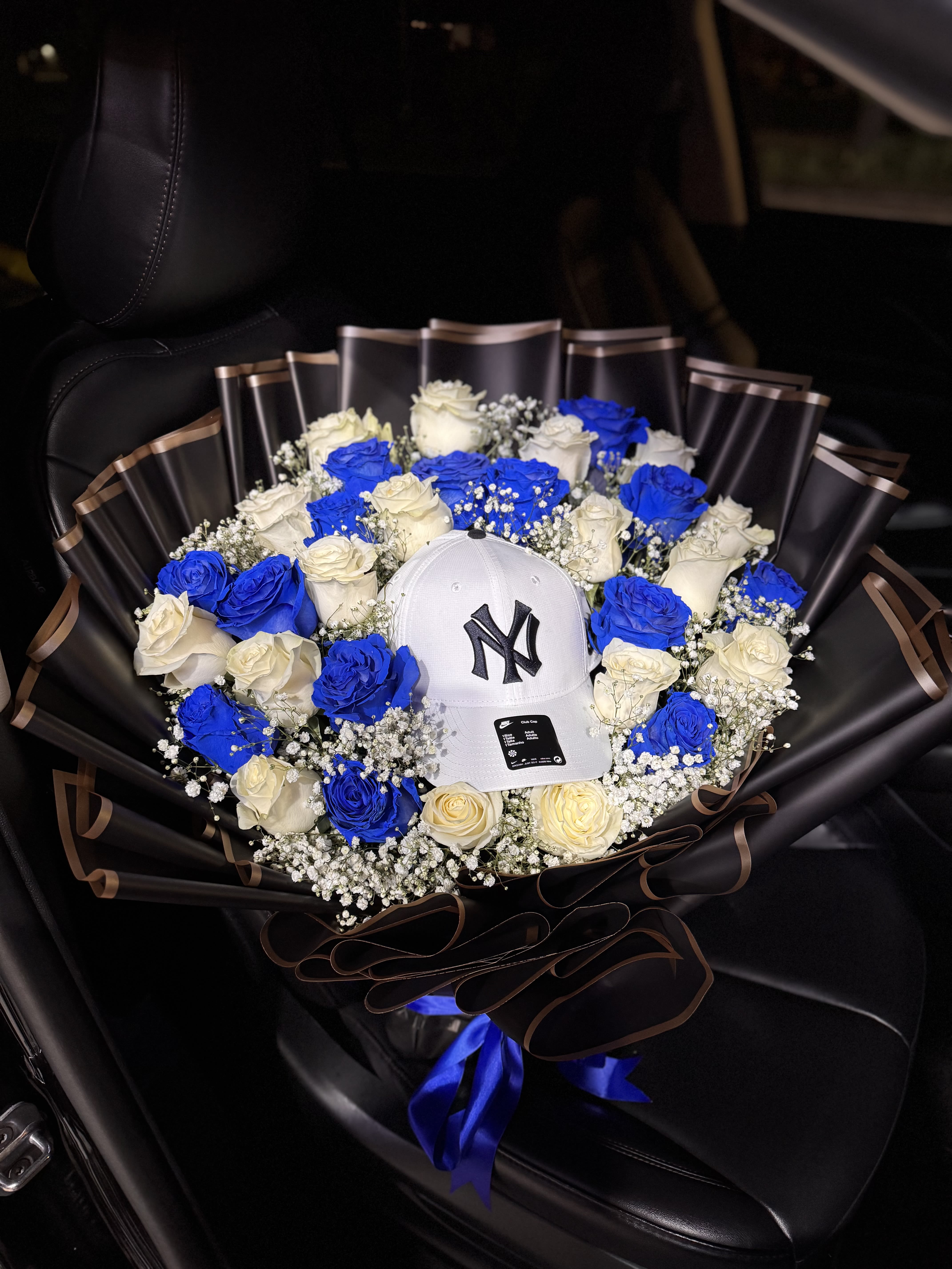 For him Blue Legacy- Custom Hat Bouquet - A bold ring of vivid blue and creamy white roses frames a crisp white baseball cap at the center-a sporty, modern flower arrangement designed as a "for him" birthday, congratulations, or romantic gift. The striking contrast of cobalt blooms against deep black wrapping paper and brown edging gives this bouquet a confident, masculine look that reads as both celebratory and thoughtfully personalized.  Tightly packed roses are softened by sprinklings of small white filler flowers (baby's breath) for airy texture, while a bright blue ribbon and a brown ribbon finish the base for a polished presentation. Wrapped in glossy black paper, the arrangement reads like a gift-in-flower form: the cap sits securely as the focal keepsake, making the whole piece feel like a stylish surprise.  Choose this for the man who loves sporty details or a unique keepsake-an eye-catching way to mark a birthday, promotion, or special milestone so he feels seen and celebrated. Available in a single custom size; handle flowers gently, keep them cool, and remove the cap before placing blooms in water if you plan to recut stems.