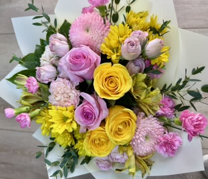 Sunrise Blush Garden - A vibrant, hand-tied bouquet blending soft romance with sunny energy. This arrangement features buttery yellow roses, blush pink roses, delicate spray roses, fluffy pink carnations, and cheerful yellow chrysanthemums, accented with fresh greenery and seasonal fillers. The soft pastel tones paired with pops of golden yellow create a warm, uplifting feel—perfect for birthdays, thank-you gifts, or brightening someone’s day.