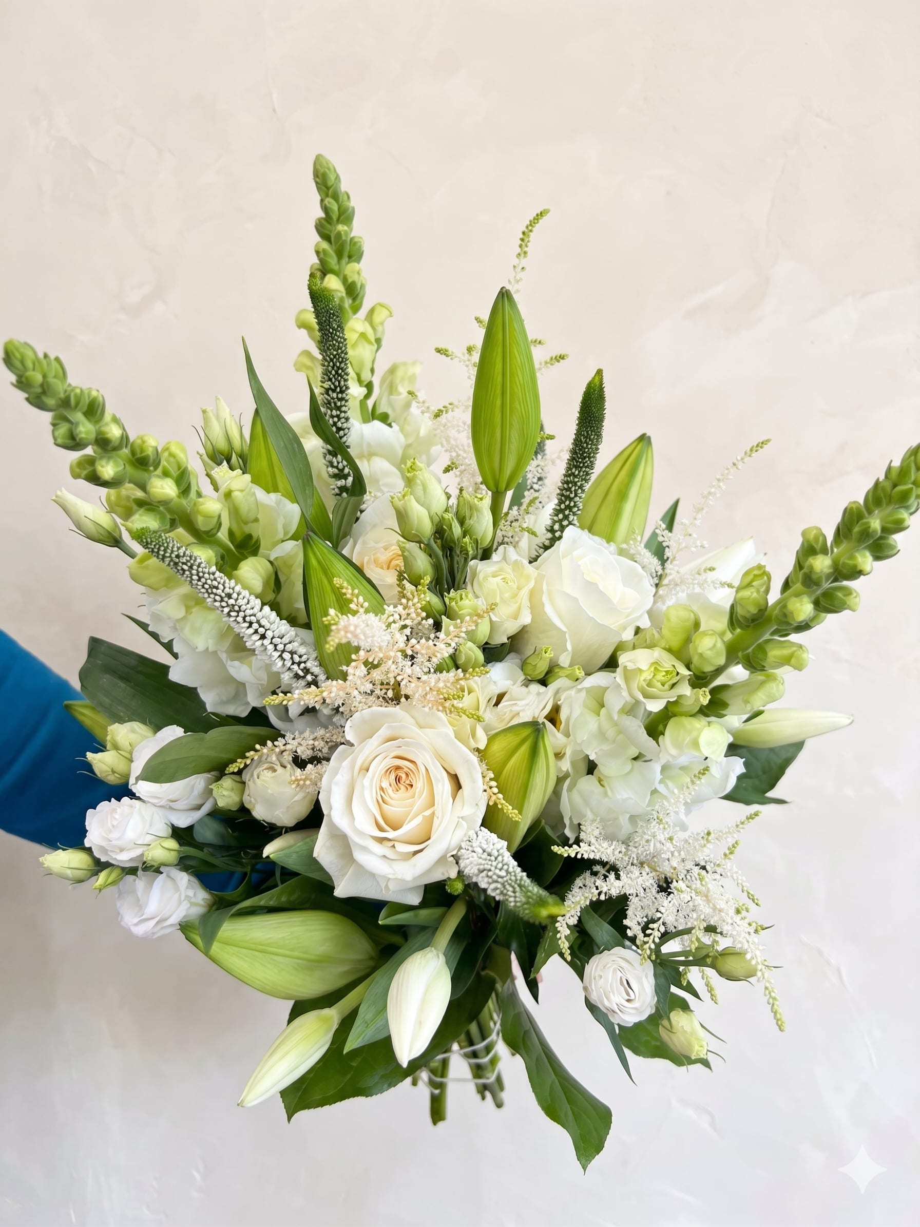 BP125 - The White Garden Bouquet - This bouquet featuring classic whites and vibrant greens. This garden-style arrangement is packed with premium blooms like white roses, lisianthus, and tall snapdragons, lilies , veronica, and astilbe.  Seasonality: Snapdragons (available spring/summer), and Astilbe (available spring/summer).