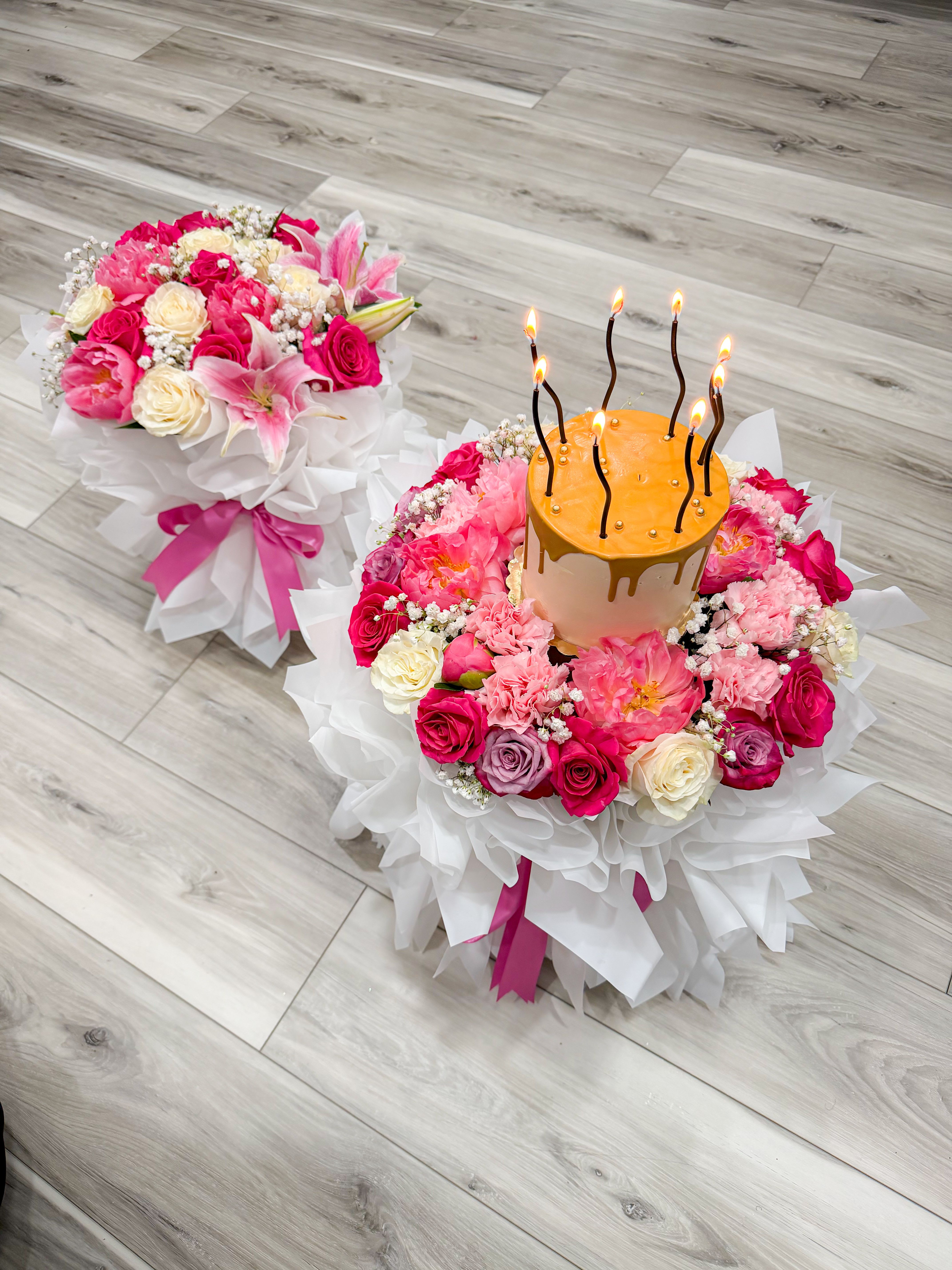 Sweet Bloom Celebration- Cake Bouquet - Two matching round bouquets wrapped in crisp white paper and tied with bright pink ribbon, each bursting with pink, white, lavender, and yellow blooms - the Sweet Bloom Celebration cake bouquet is built around a small gold‑frosted cake topped with lit birthday candles. This birthday bouquet (also perfect for anniversaries or congratulations) reads unmistakably festive and feminine: the candlelit cake creates an immediate celebratory focal point so the person you're honoring will feel seen and special the moment it arrives.  Dense, garden‑style clusters of roses, carnations, lilies, daisies and delicate baby's breath form a lush, rounded presentation around the cake, with cream and blush tones balancing vibrant hot‑pink highlights. The set includes two full arrangements, each wrapped in layered white paper and finished with vivid pink ribbon for a polished gift‑set look; metallic bead accents on the cake add a touch of sparkle. Ideal for gifting at a birthday party, anniversary breakfast, or surprise delivery, this duo arrives ready to display and enjoy - cake, candles, and flowers combined for an unforgettable celebratory moment.