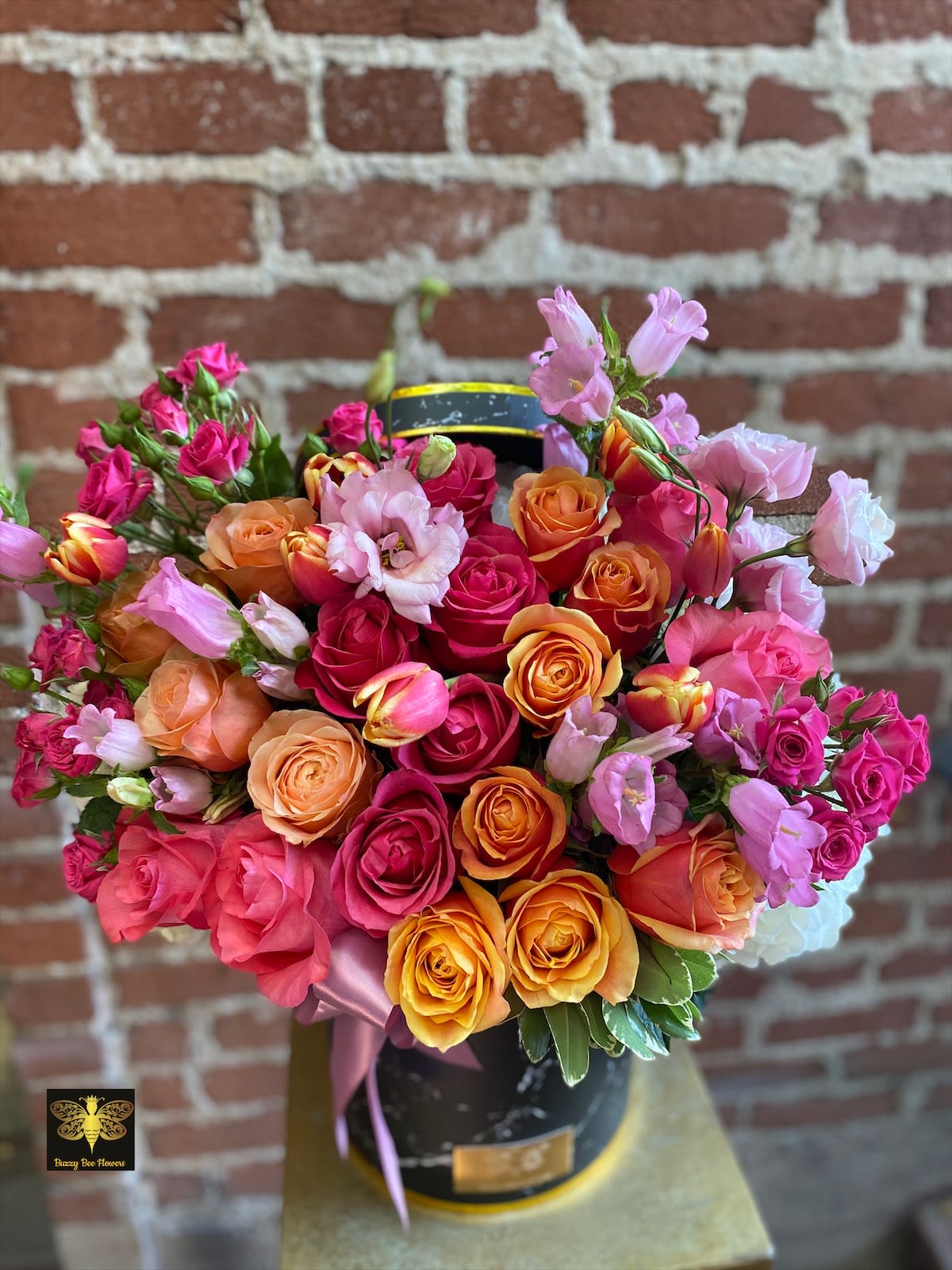 Bouquet of pink, orange, and yellow roses with light purple blooms in a black hatbox